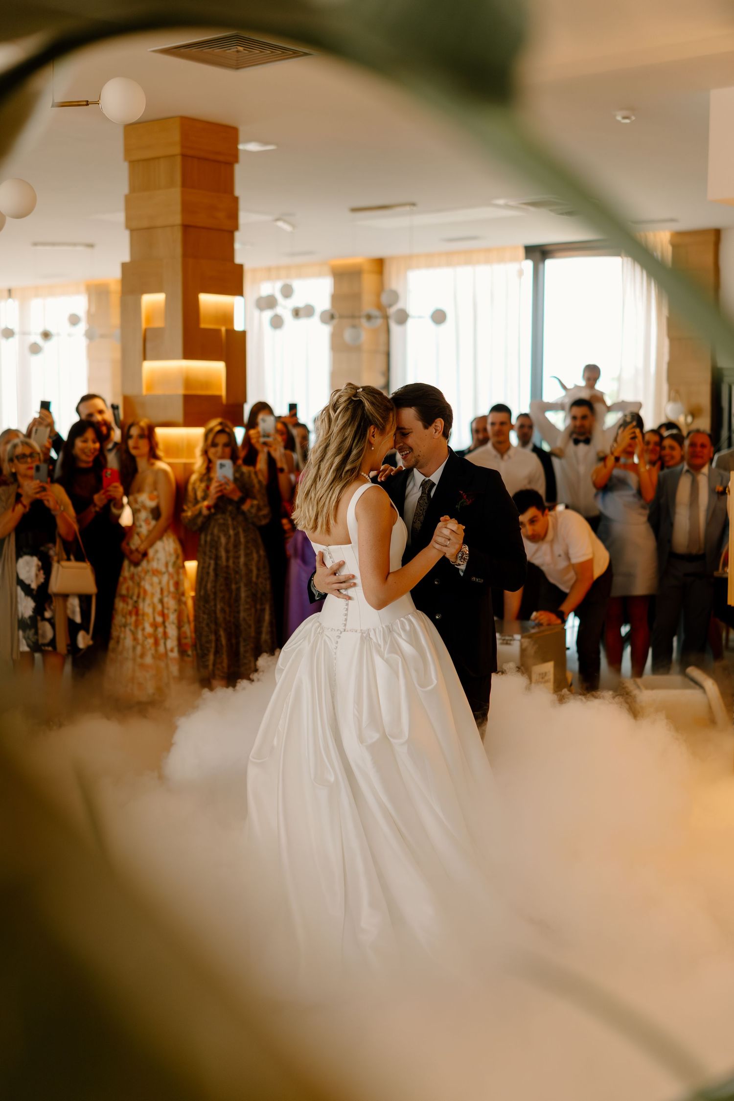 Bride and groom share their first dance surrounded by fog and wedding guests in an elegant venue.