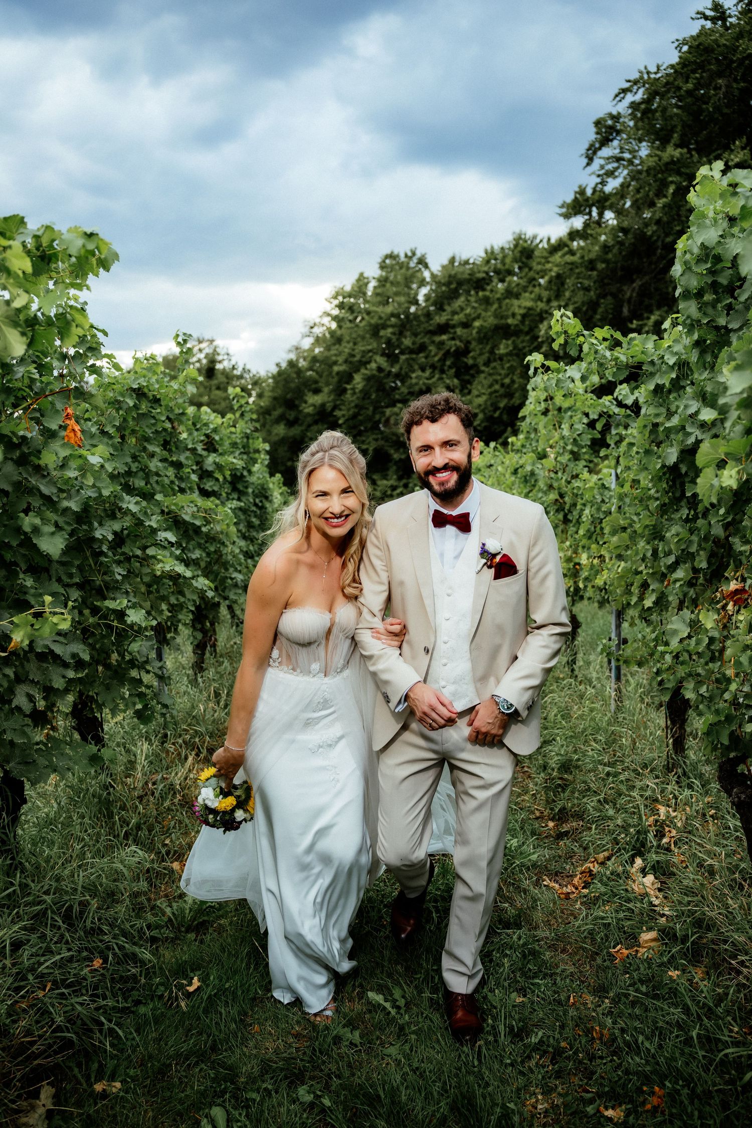Smiling bride and groom walking through lush vineyard rows, bride holding colorful bouquet, groom in beige suit with burgundy bow tie.