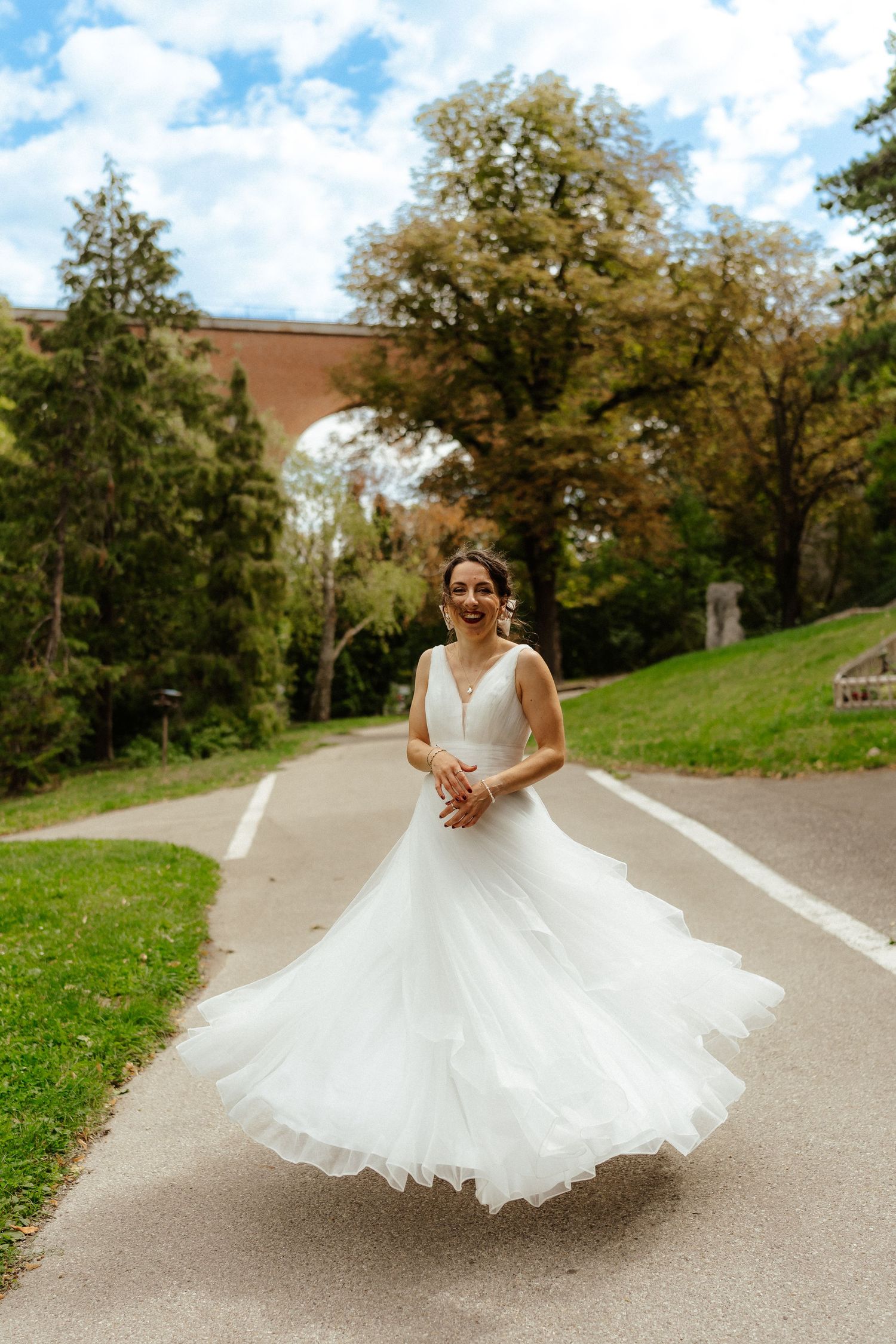 Joyful bride in a flowing white tulle gown twirling on a park path with lush trees and an arch bridge behind her.