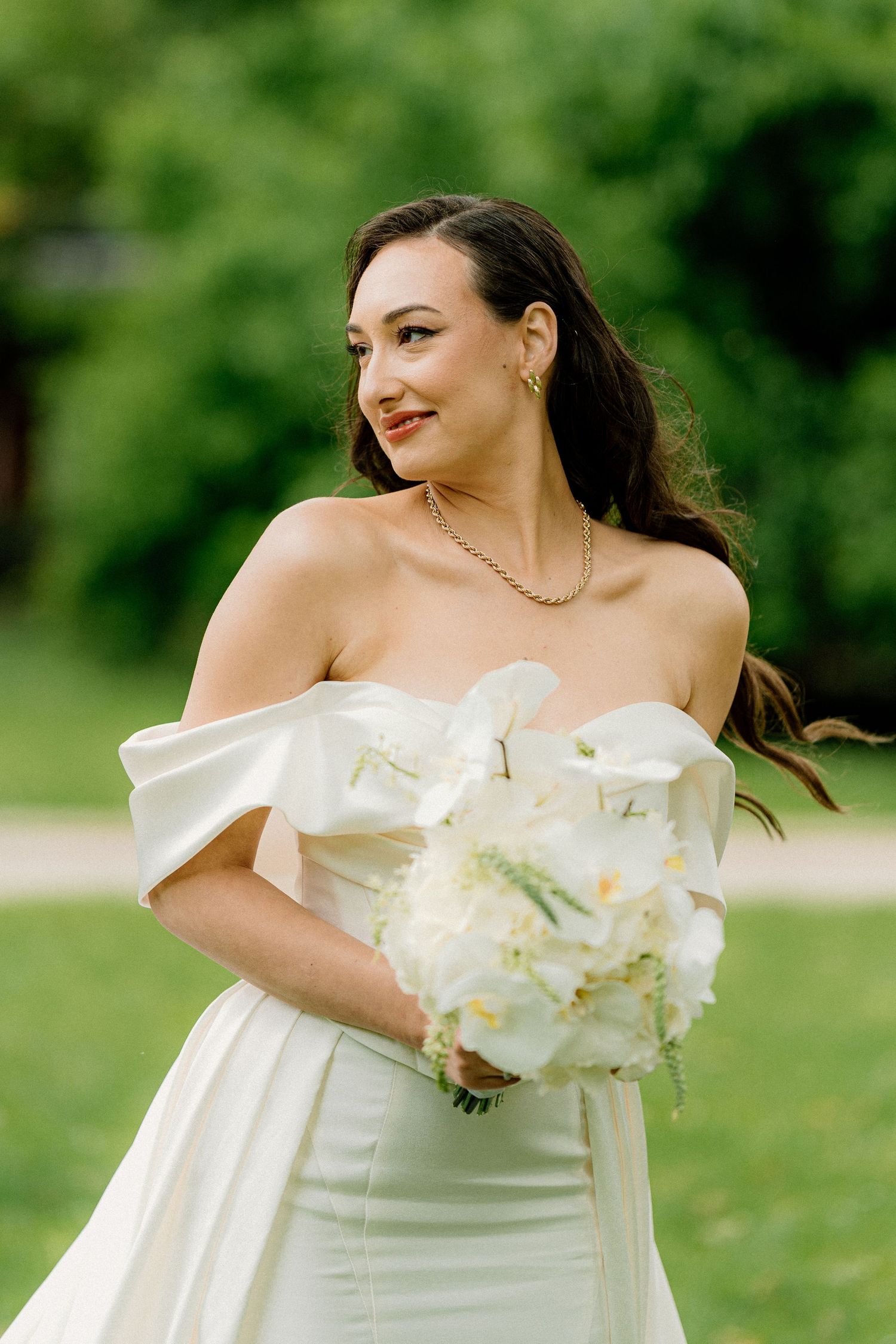 Bride in off-shoulder gown poses with white orchid bouquet against green background.