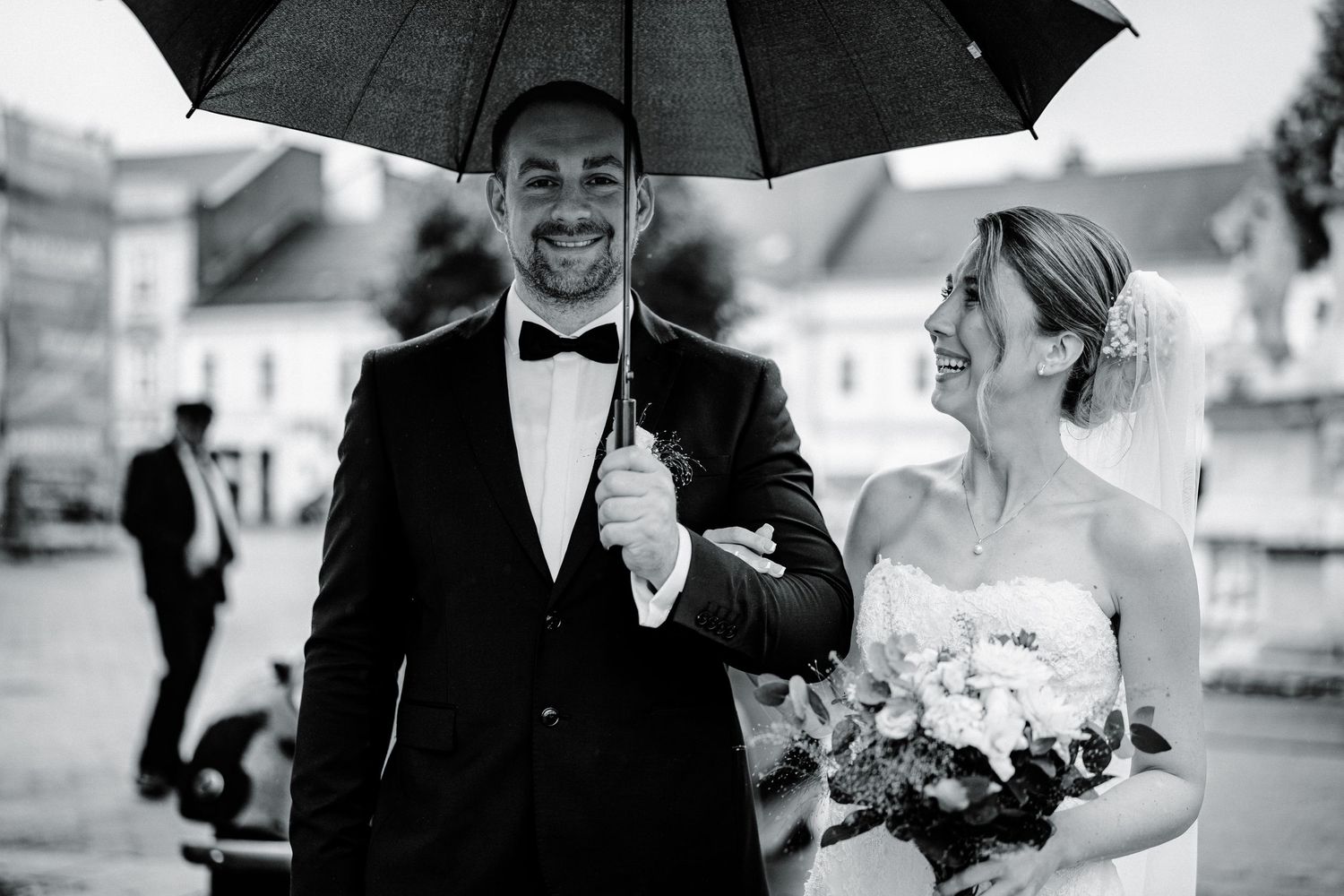 A joyful wedding couple shares an umbrella while walking together outdoors.