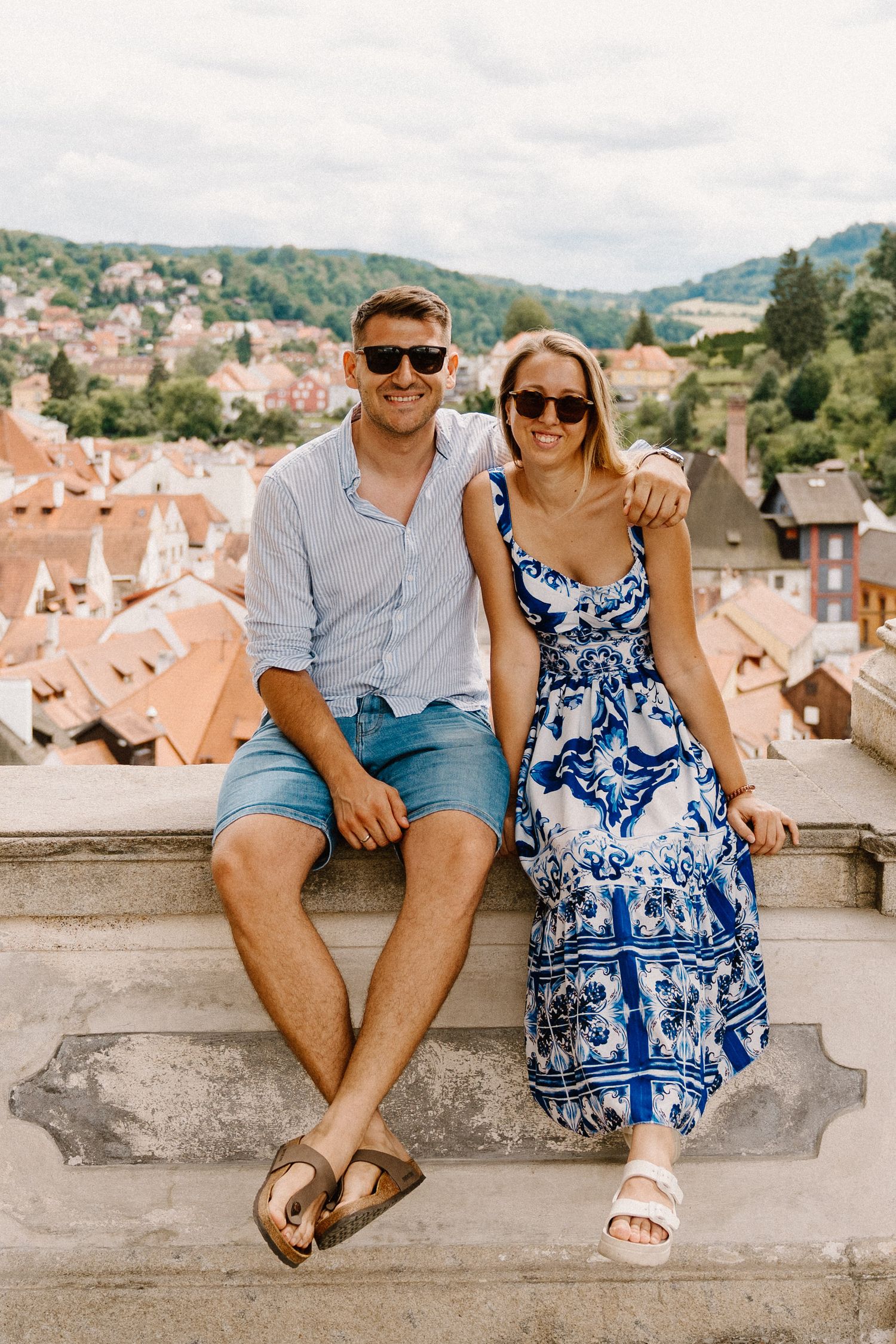 A couple sits together on a stone wall overlooking European rooftops on a sunny day.