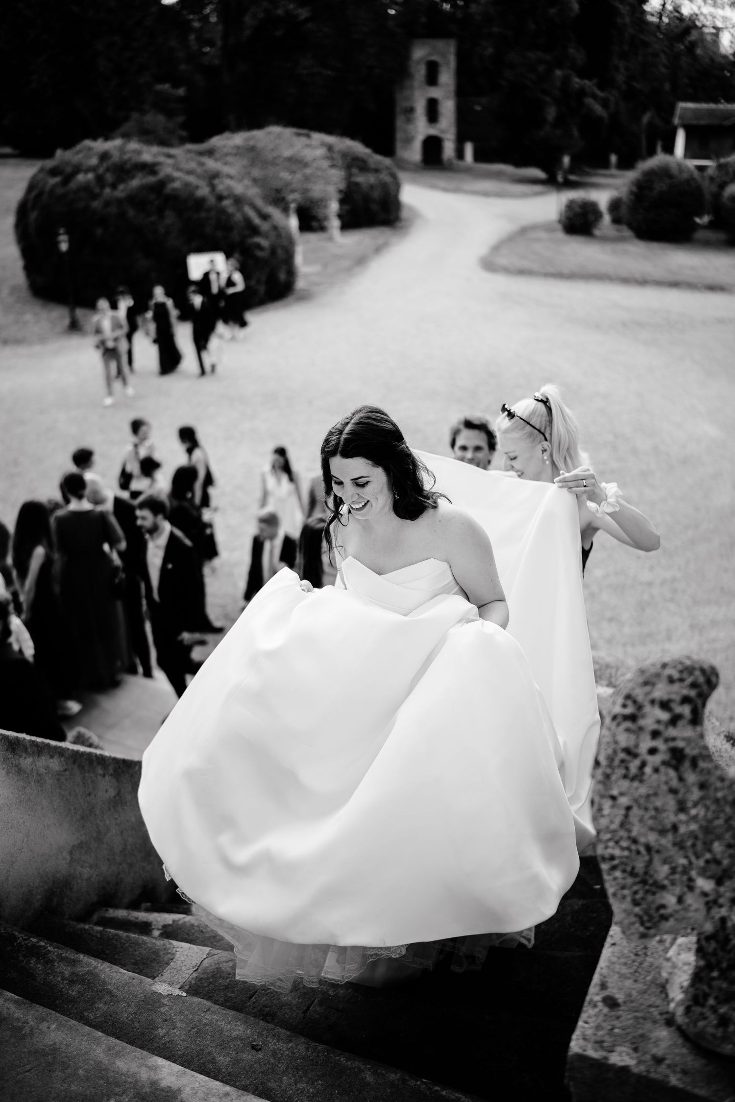 Black and white photo of a bride ascending stone steps in a flowing gown, assisted by a bridesmaid at an elegant venue.