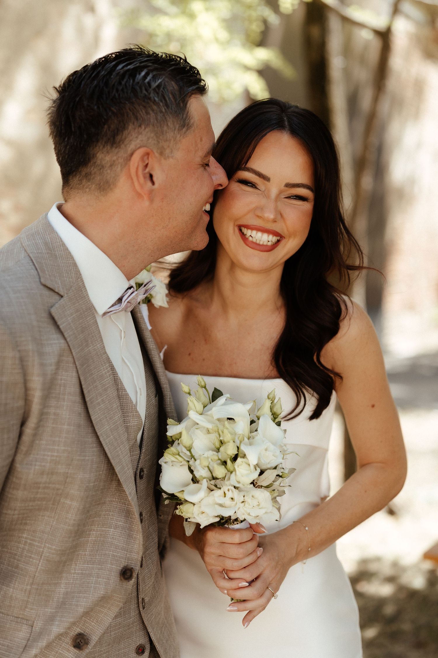 Smiling bride in white strapless gown holds white bouquet as groom in tan suit whispers in her ear outdoors.