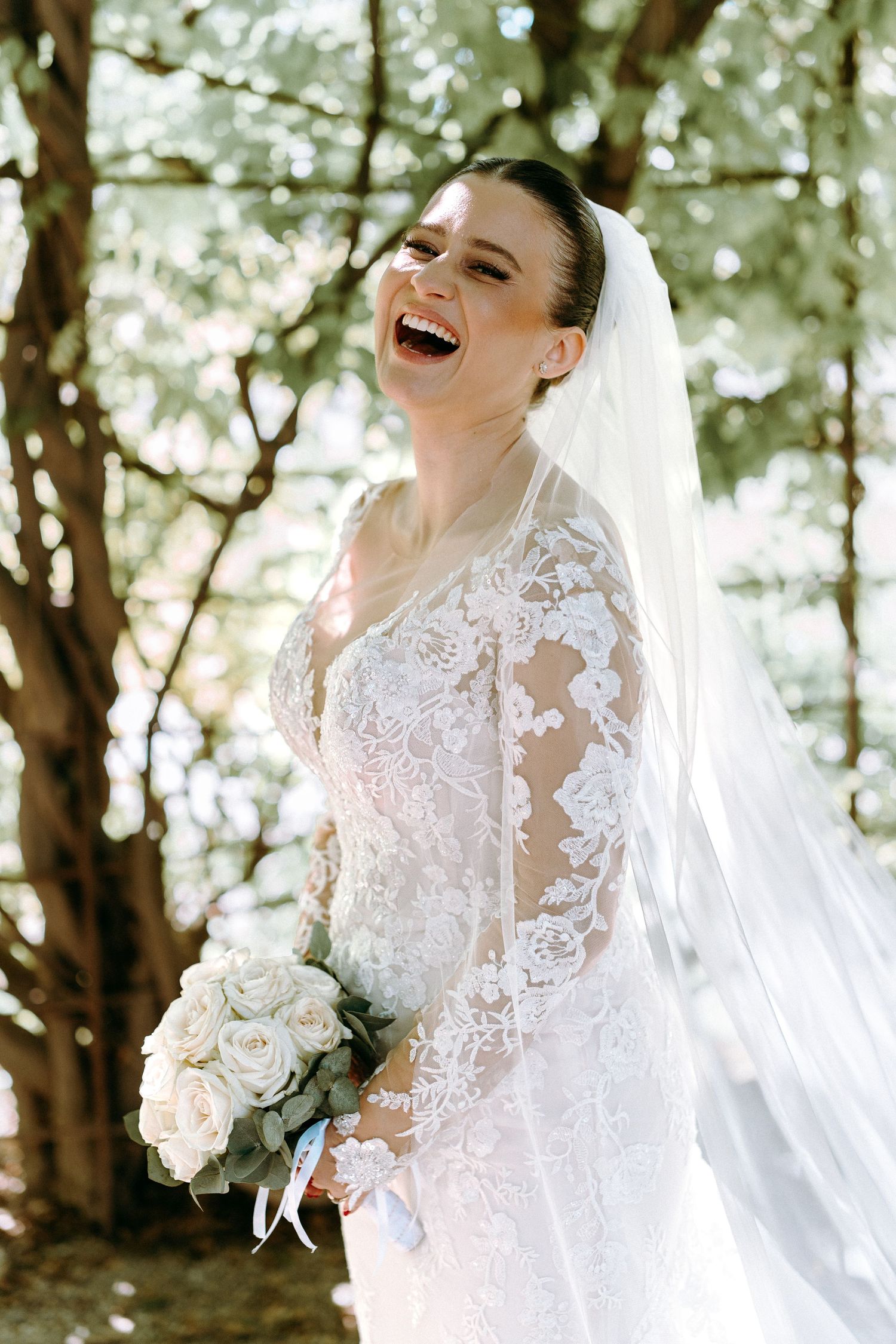 A joyful bride in a lace wedding dress laughing while holding a white bouquet outdoors.
