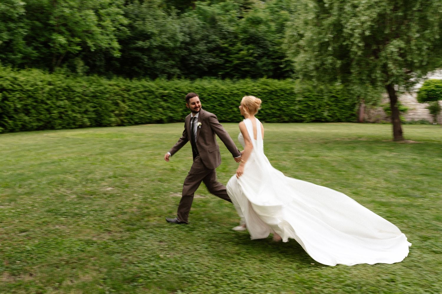 Bride in flowing white gown and groom in brown suit walk hand in hand across lush green lawn at outdoor wedding.