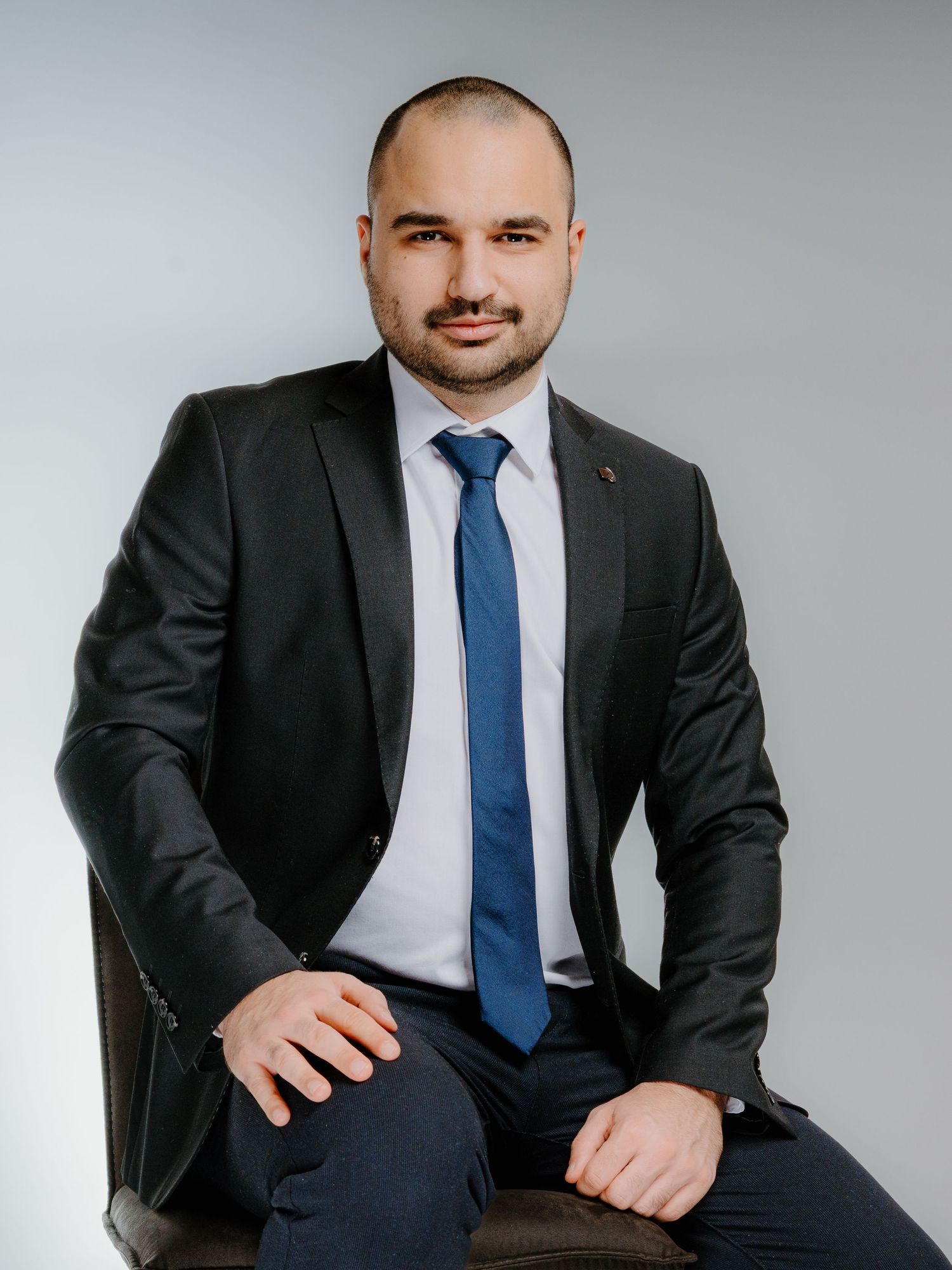 Professional businessman in dark suit and blue tie sitting confidently in a chair against a light gray background.