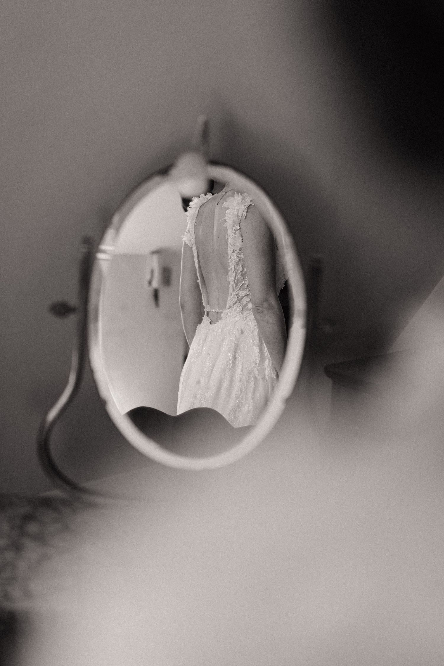 Black and white photo of a wedding dress reflected in a small oval mirror, showcasing delicate lace details.