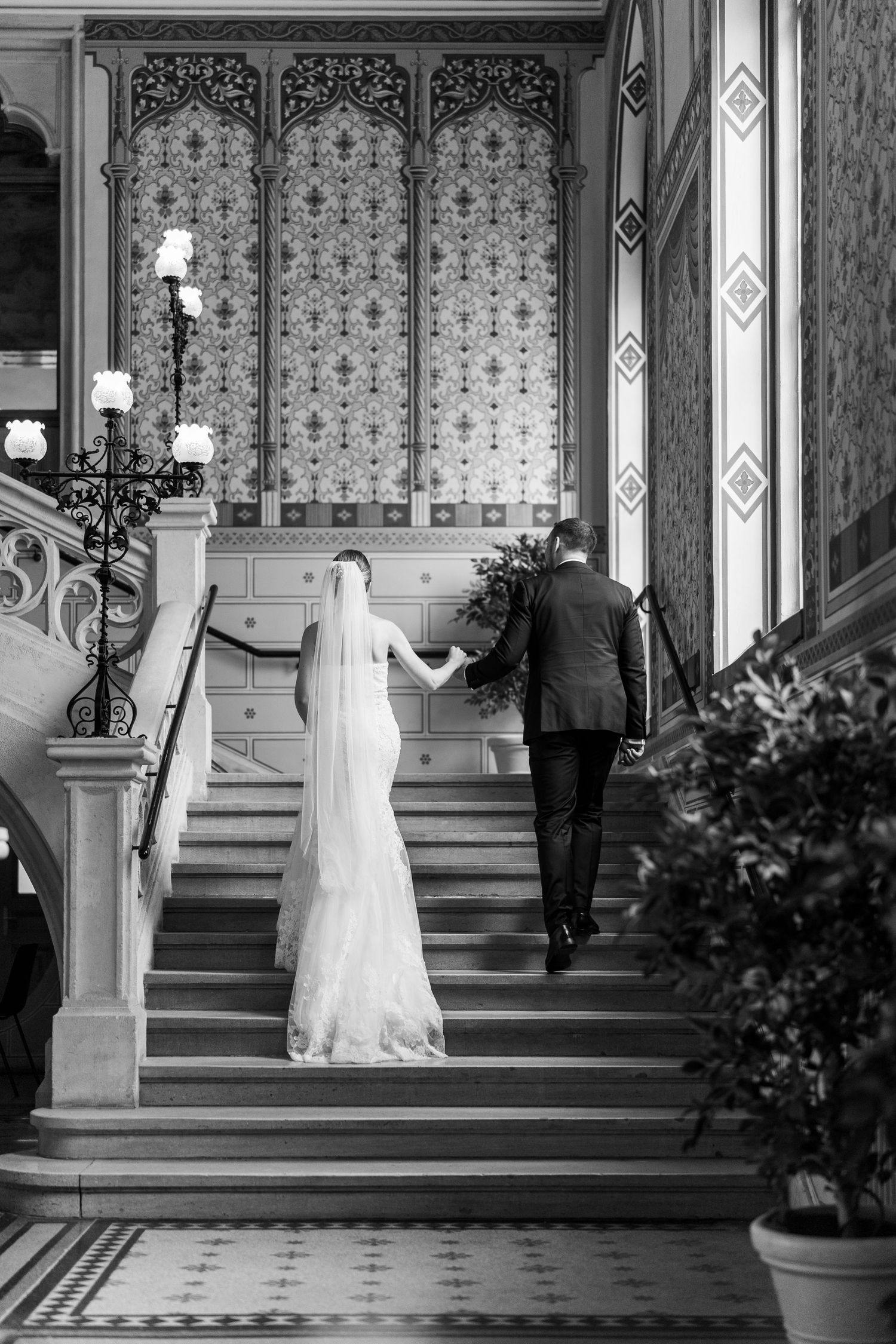 A couple ascends an elegant staircase with ornate wallpaper and architectural details.