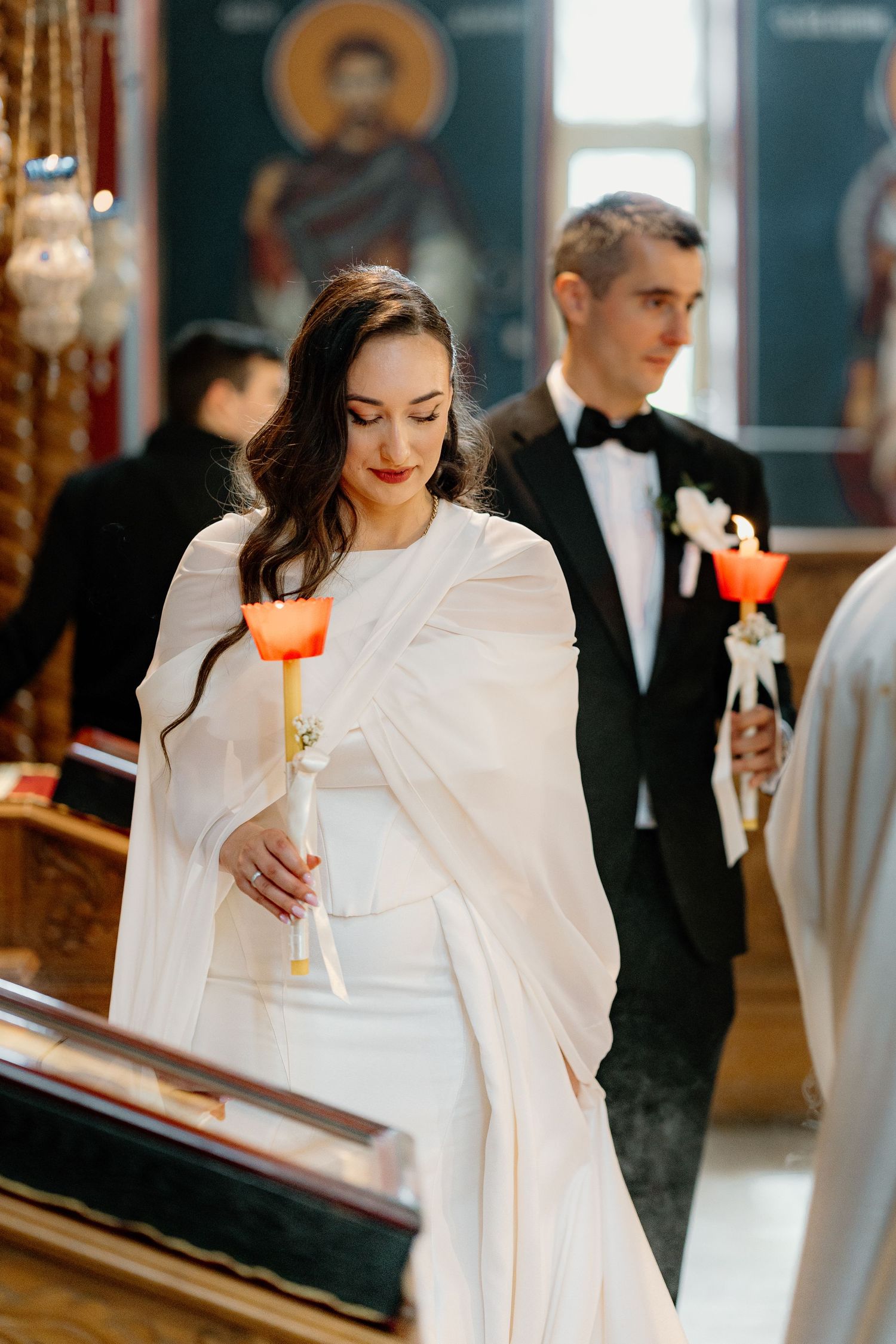 Bride in elegant white dress holds a lit candle during an indoor ceremony.
