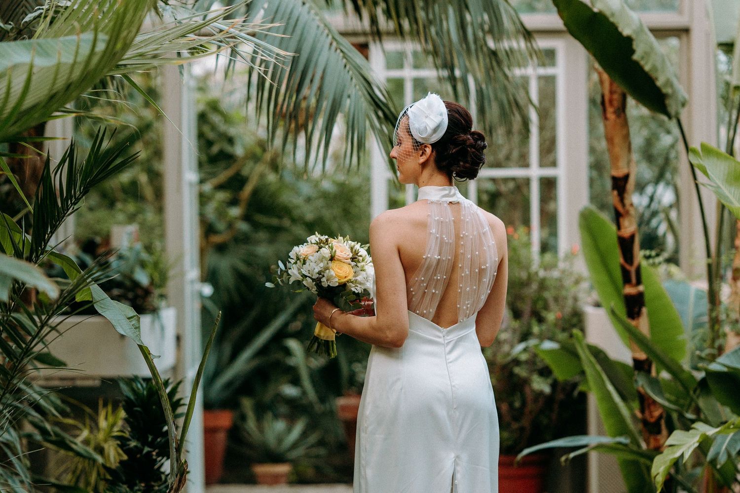 Elegant bride in backless white dress holds bouquet in greenhouse setting.