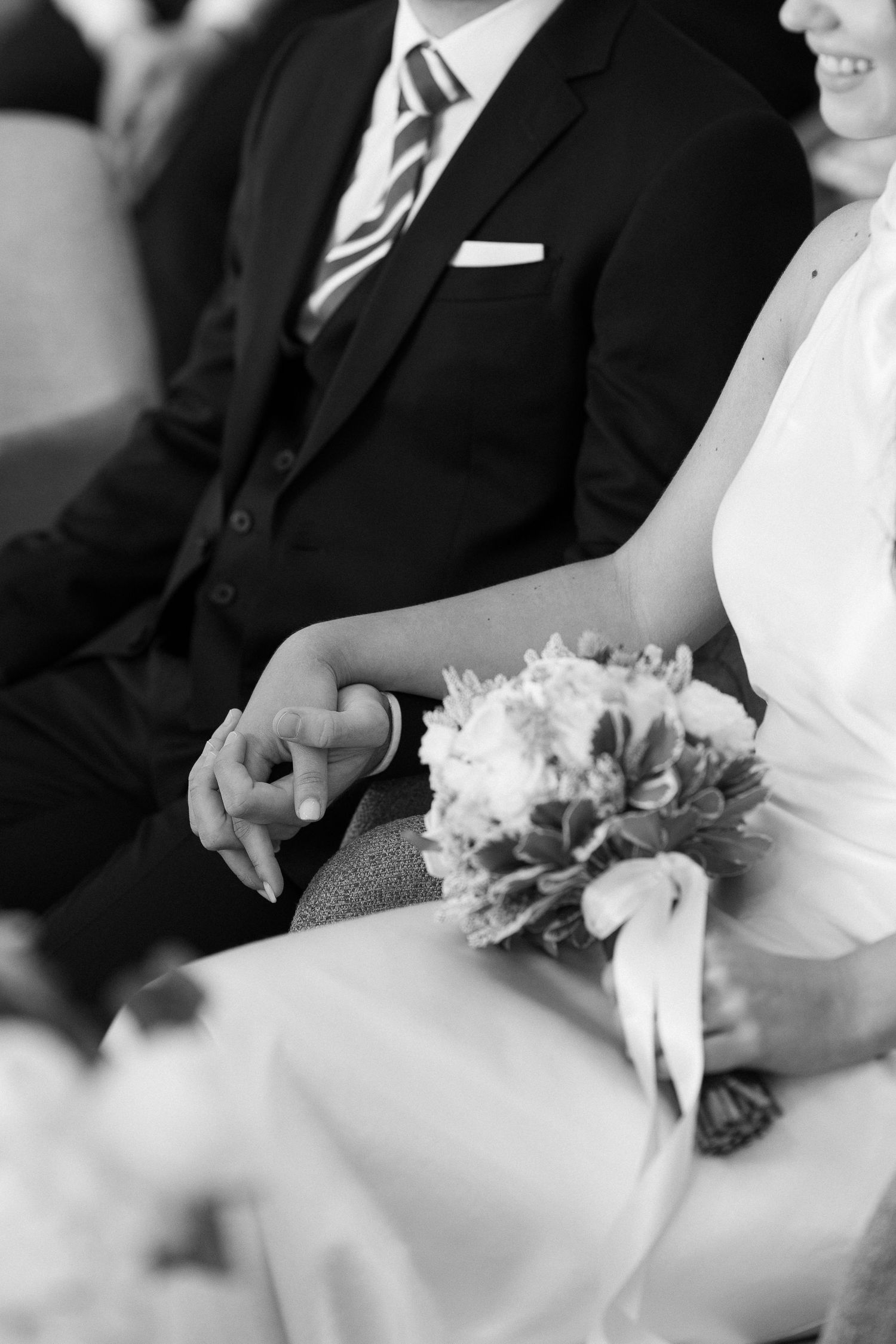 A black and white detail shot of a couple's hands and wedding bouquet during the ceremony.