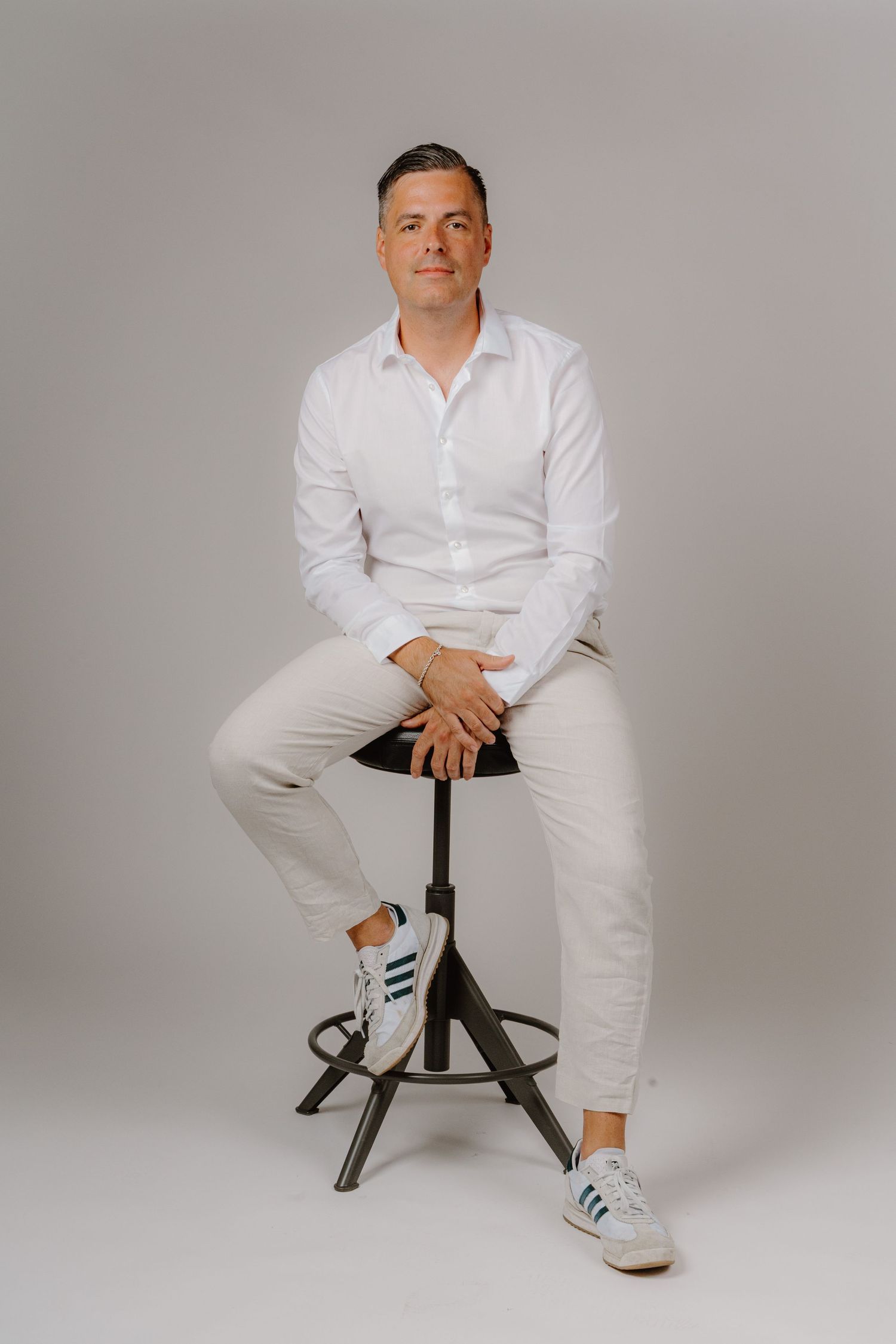 A man in a white shirt and beige pants sits on a stool wearing Adidas sneakers against a gray background.