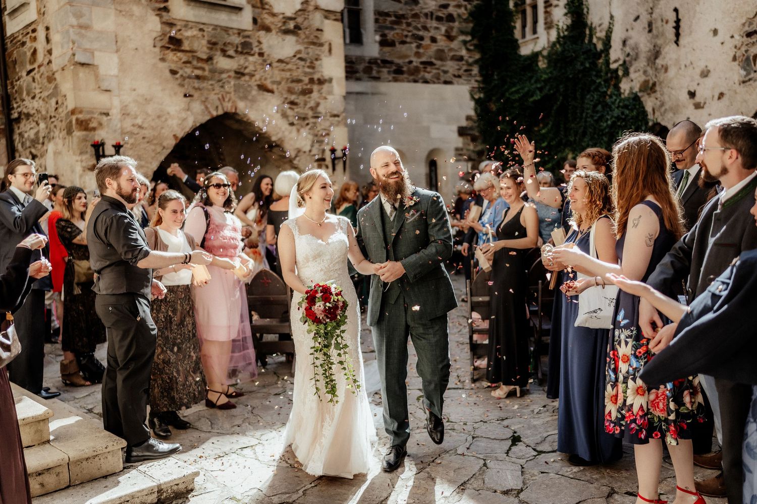 Bride in a white gown holding a red bouquet walks hand in hand with her groom as wedding guests cheer in a stone castle courtyard.