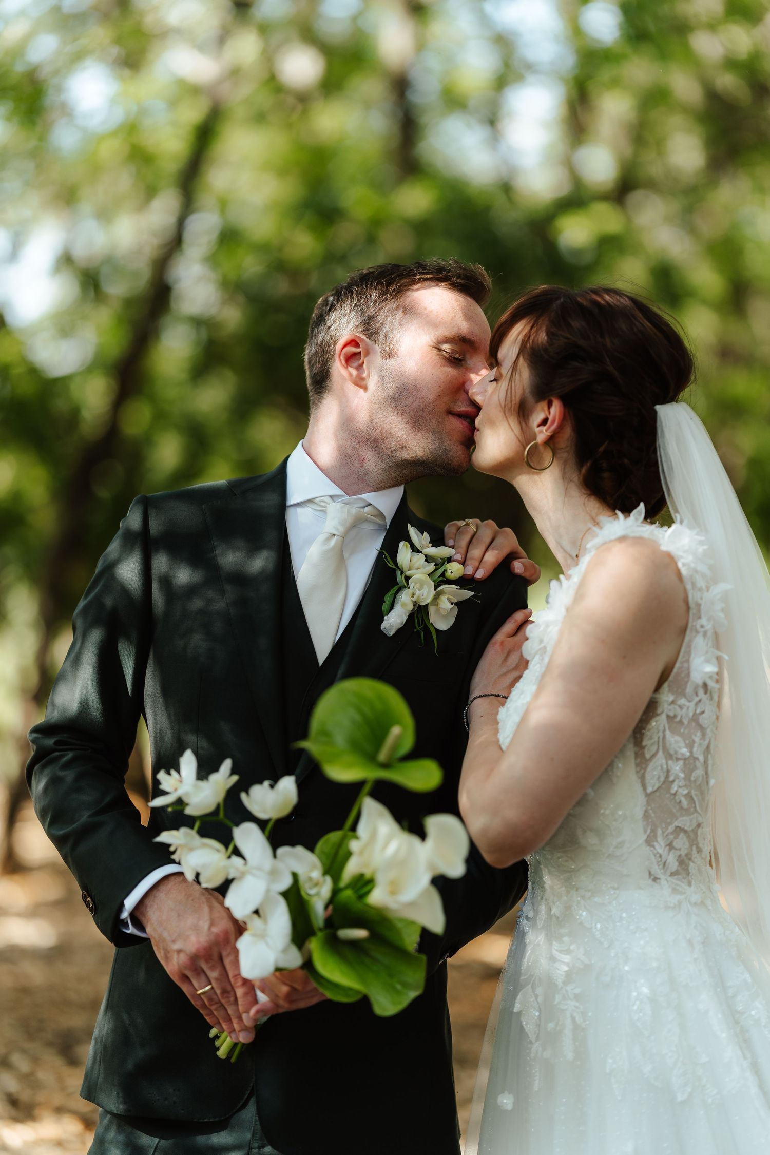 Bride and groom share a romantic kiss outdoors, groom holding white floral bouquet, surrounded by lush green trees.