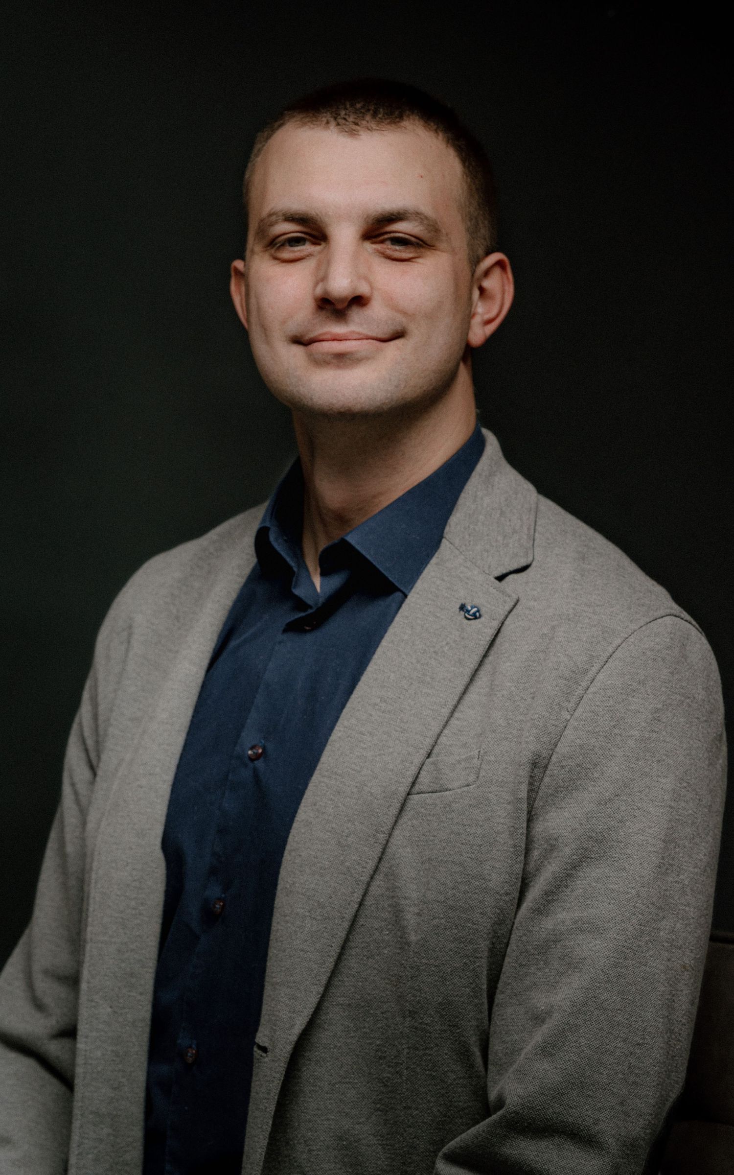 Professional man in gray blazer and navy shirt smiling confidently against dark background.