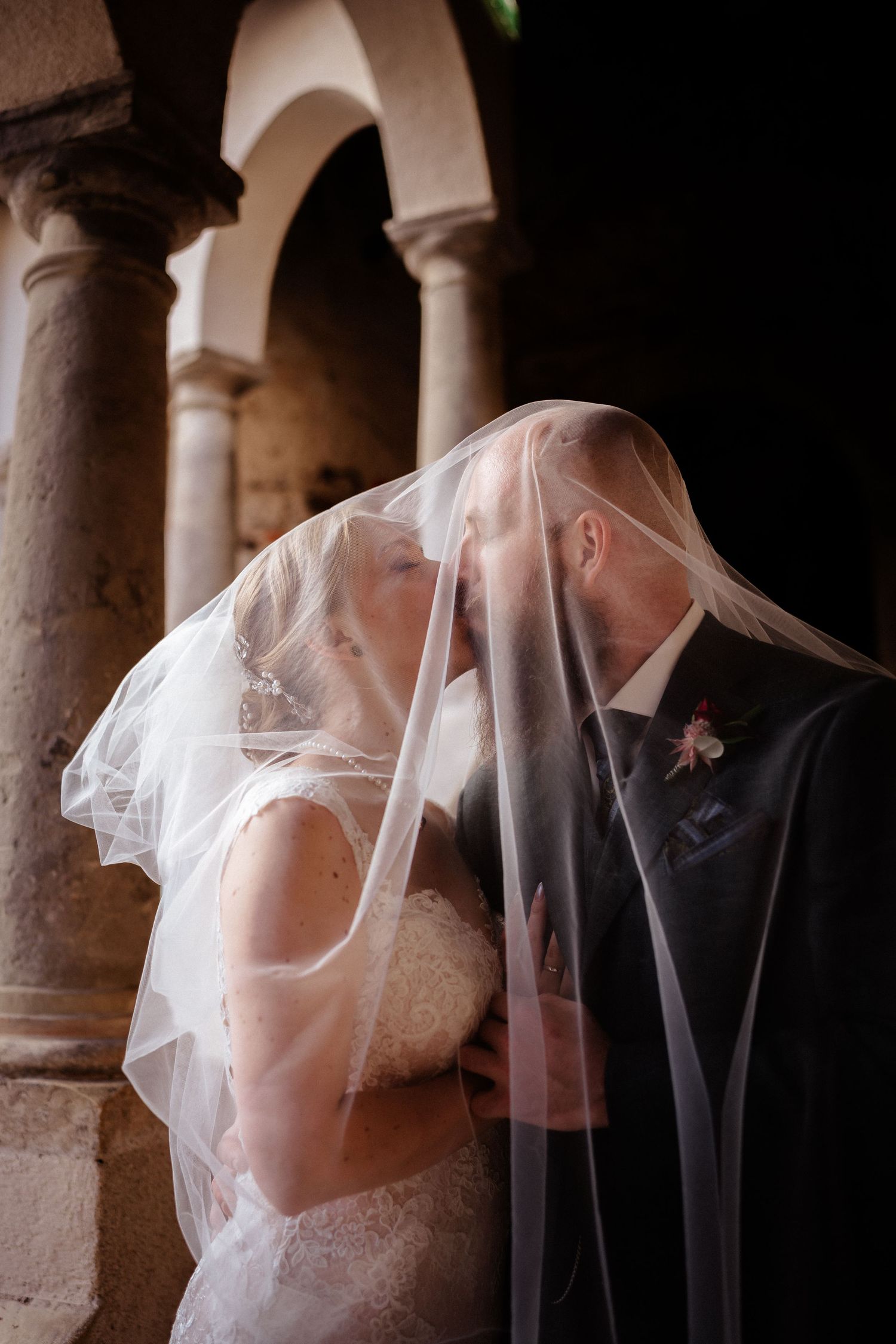 Bride and groom kissing under a flowing white veil beside stone columns in a romantic wedding portrait.