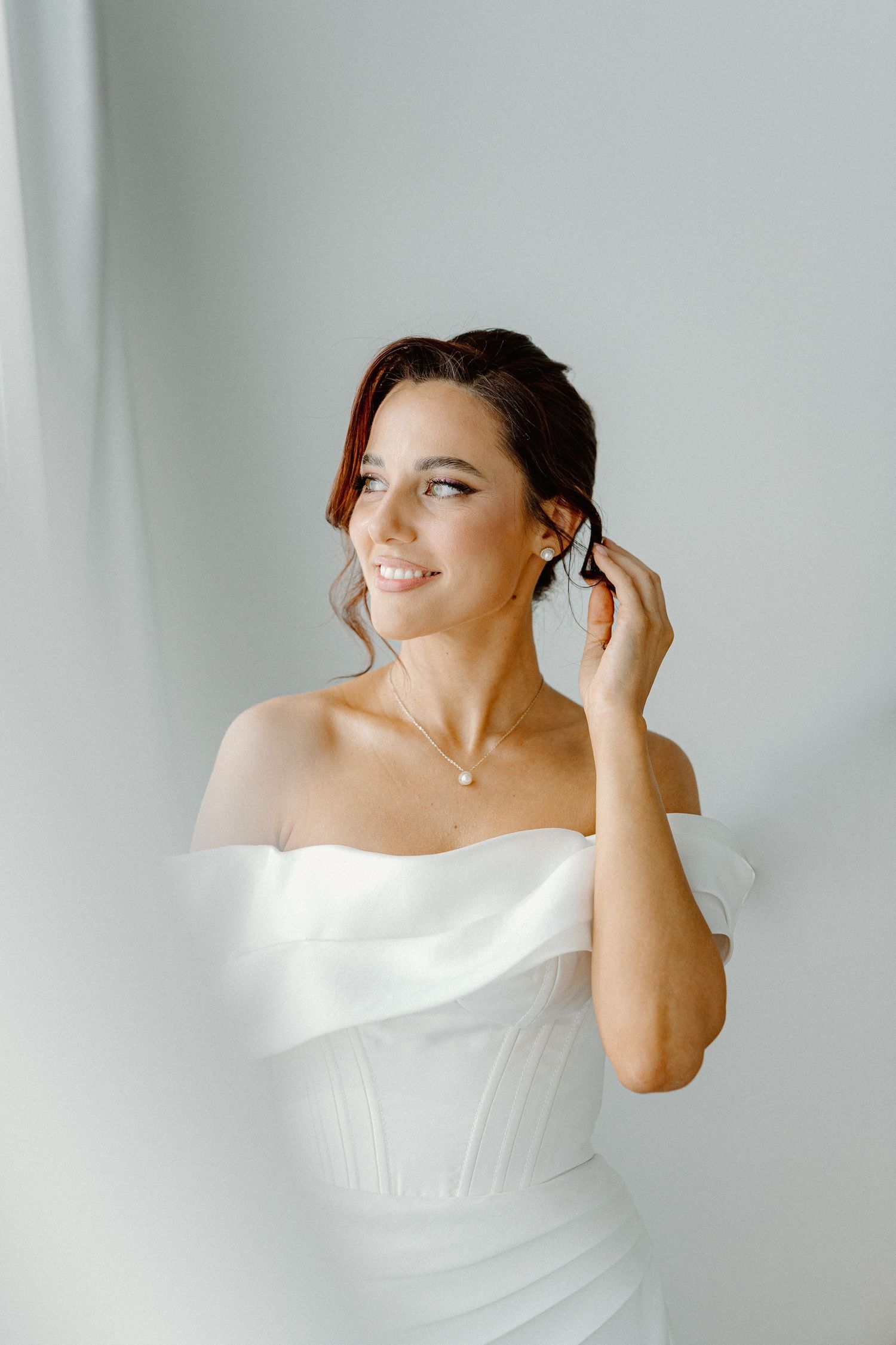 Elegant bride in off-shoulder white dress adjusting earring against light background.