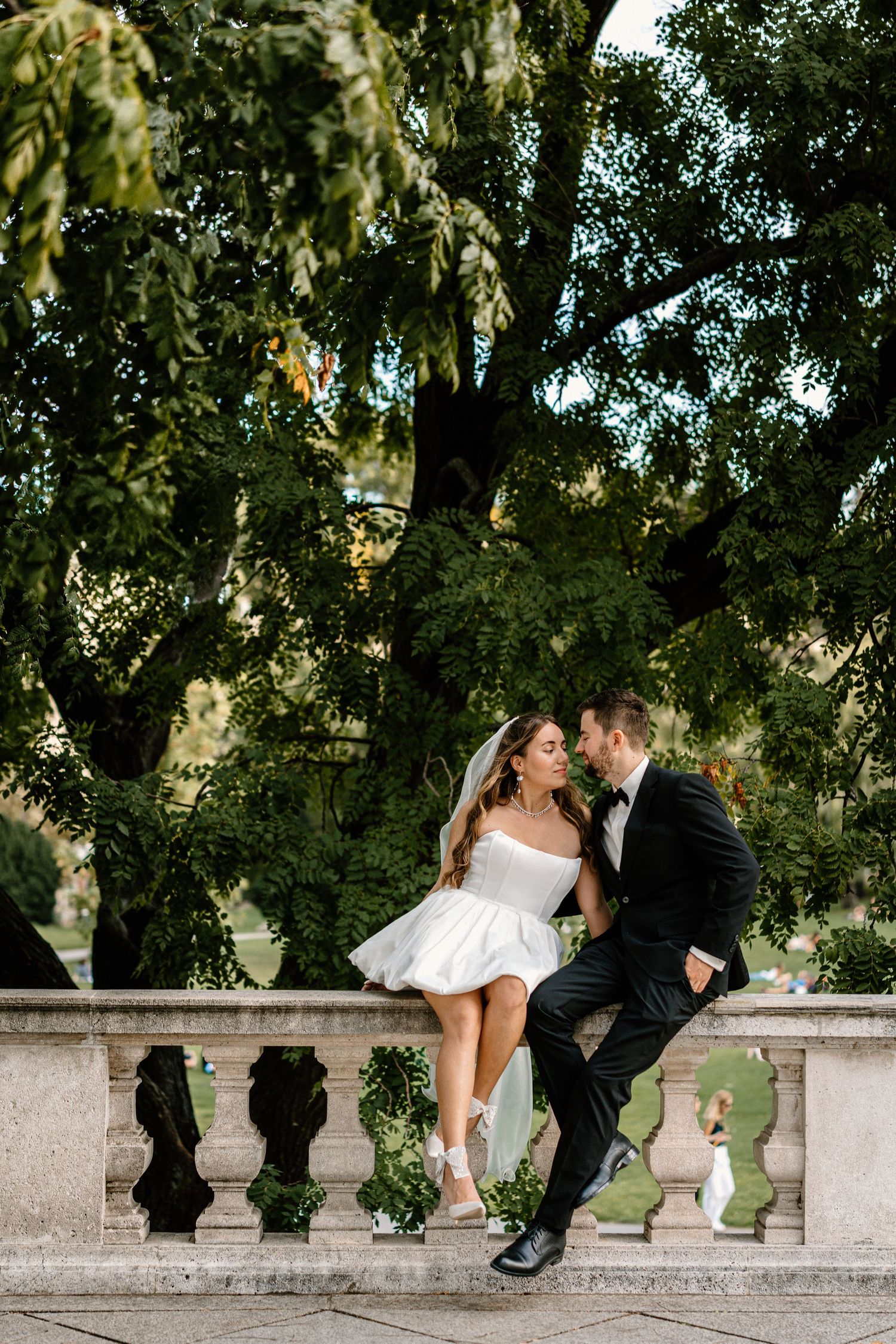 A bride and groom share a romantic kiss while sitting on a stone balustrade surrounded by lush greenery.
