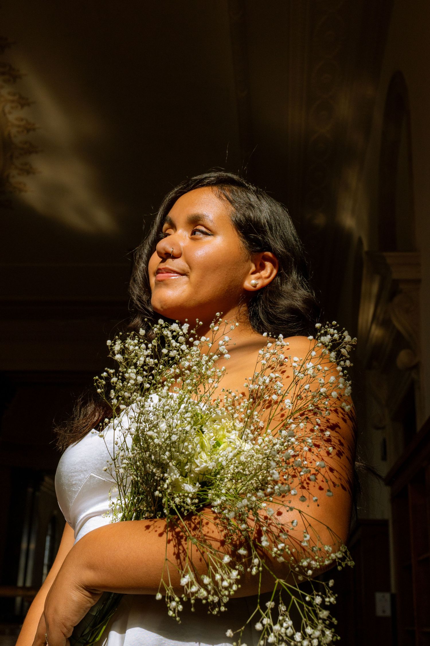 Young woman with dark hair holds white wildflower bouquet in warm sunlight, looking upward peacefully.