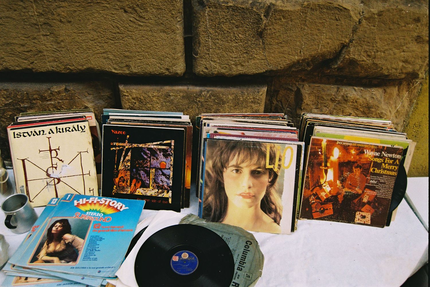 Vintage vinyl record collection displayed on table at outdoor flea market against brick wall.