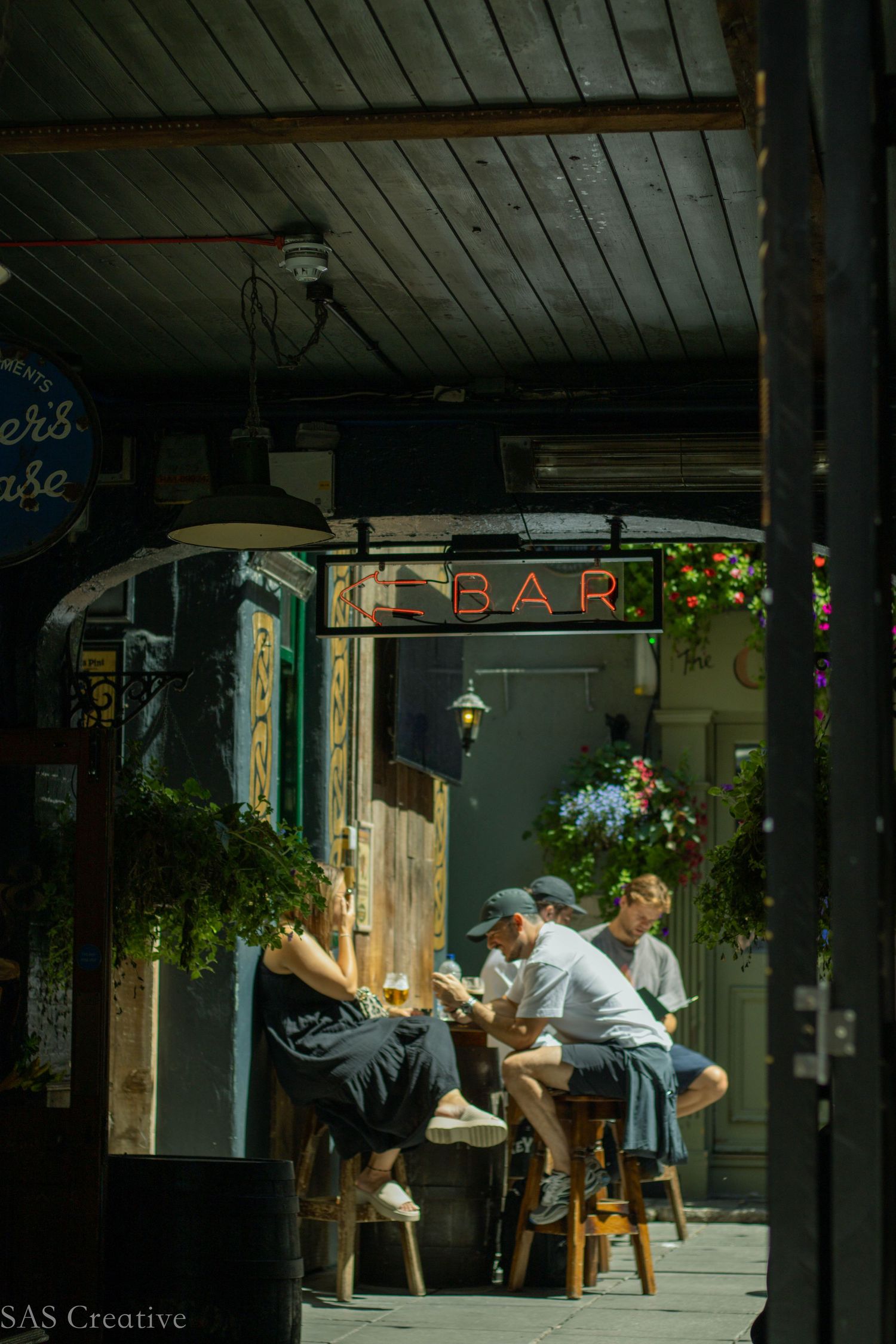 People sitting at outdoor bar stools beneath a glowing BAR neon sign at a charming pub.