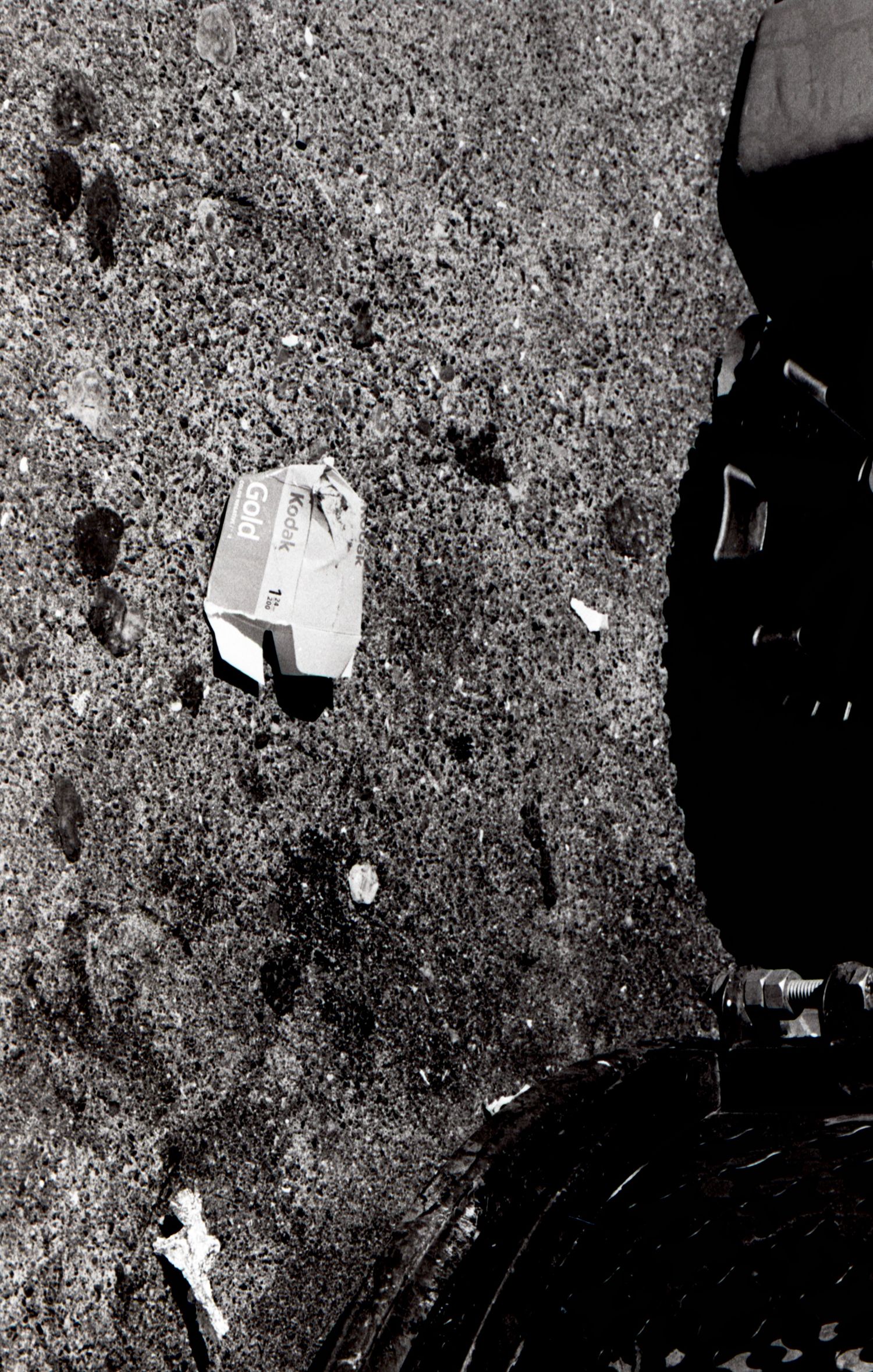 Black and white photo of a discarded Coca-Cola box on gravel ground near a bicycle wheel.