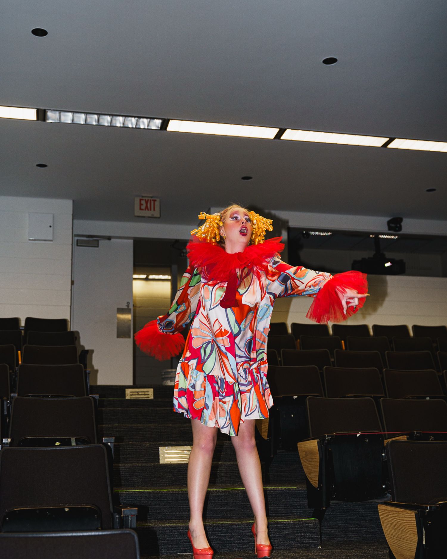 Performer in colorful printed dress with red feather collar and orange wig poses in an empty auditorium.