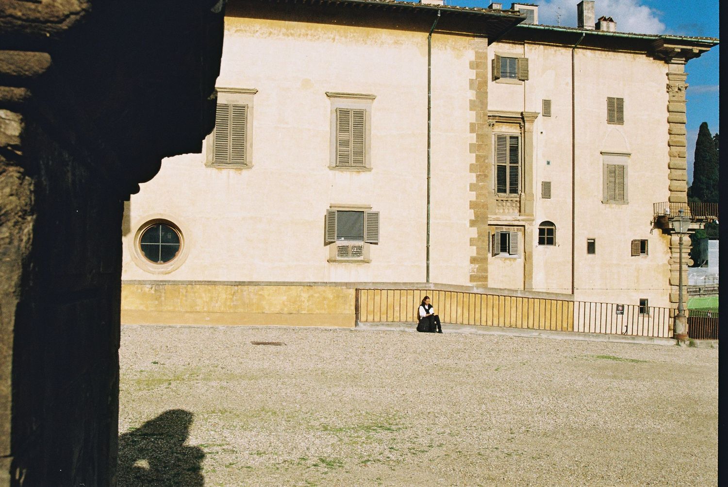 Person sitting alone in courtyard of historic Italian villa with white facade and shuttered windows.