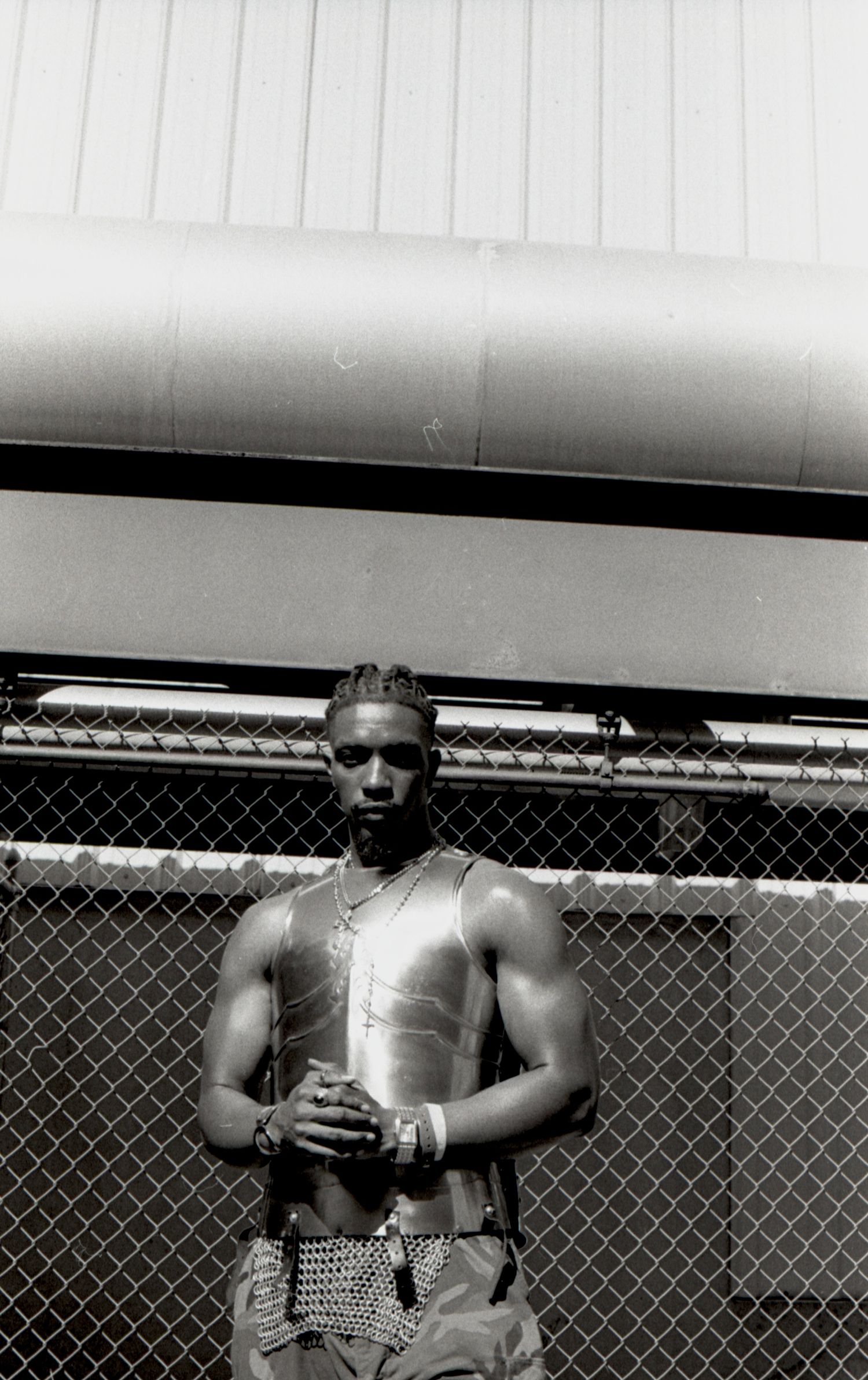 Black and white portrait of a muscular man in a metallic vest standing by a chain-link fence.