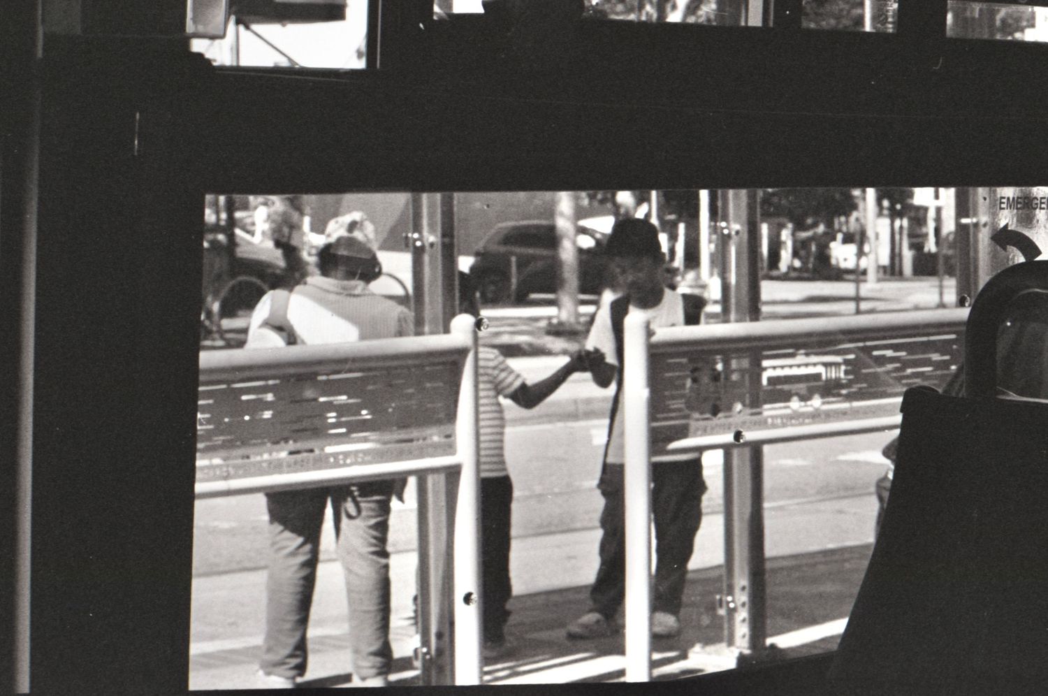 Black and white photo of people standing at a bus stop viewed through a window reflection.