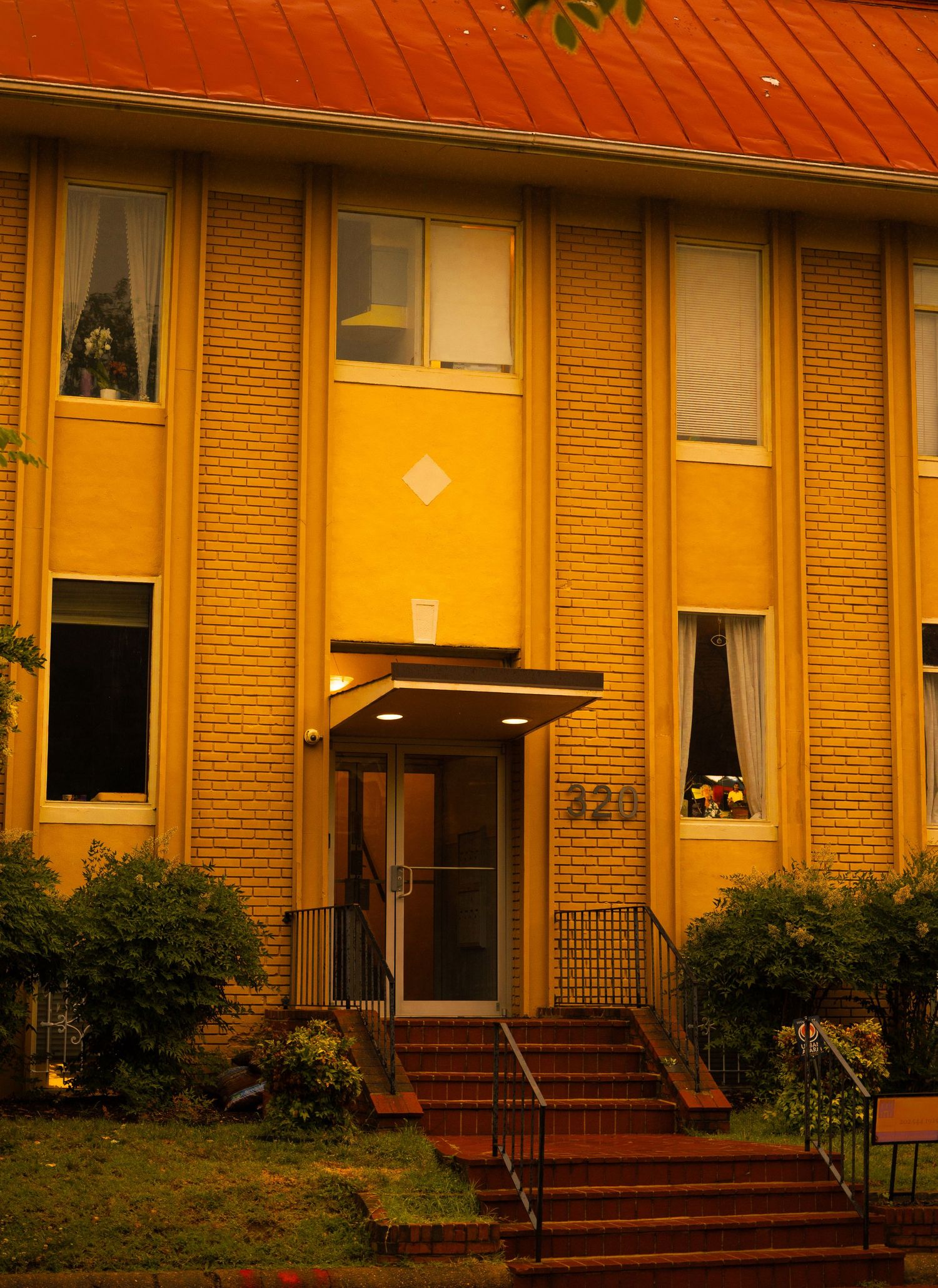 Brick building exterior with number 320, featuring tall windows, red roof, and illuminated entrance steps at dusk.