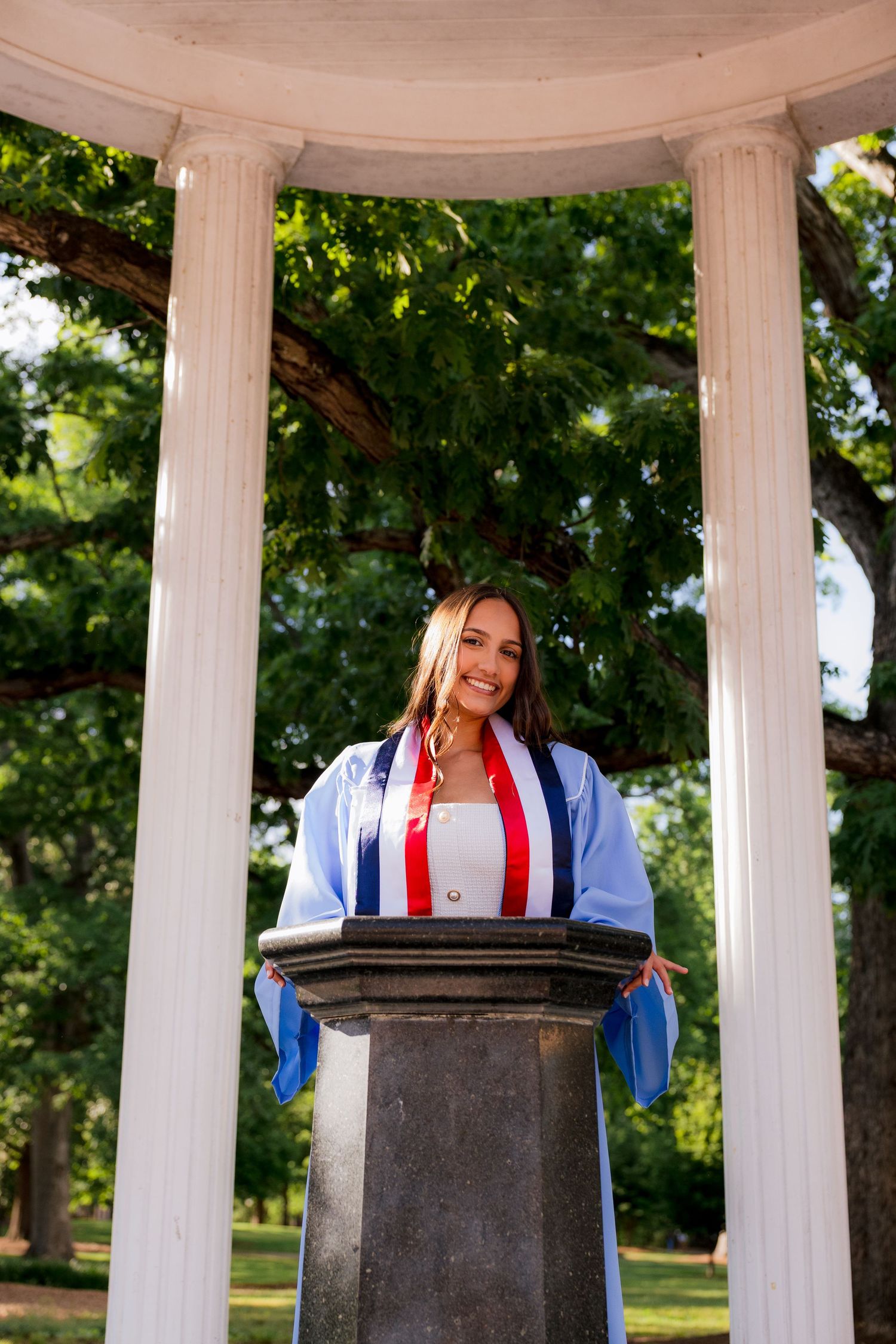 Smiling graduate in blue cap and gown stands at stone podium under white columns in a park setting.