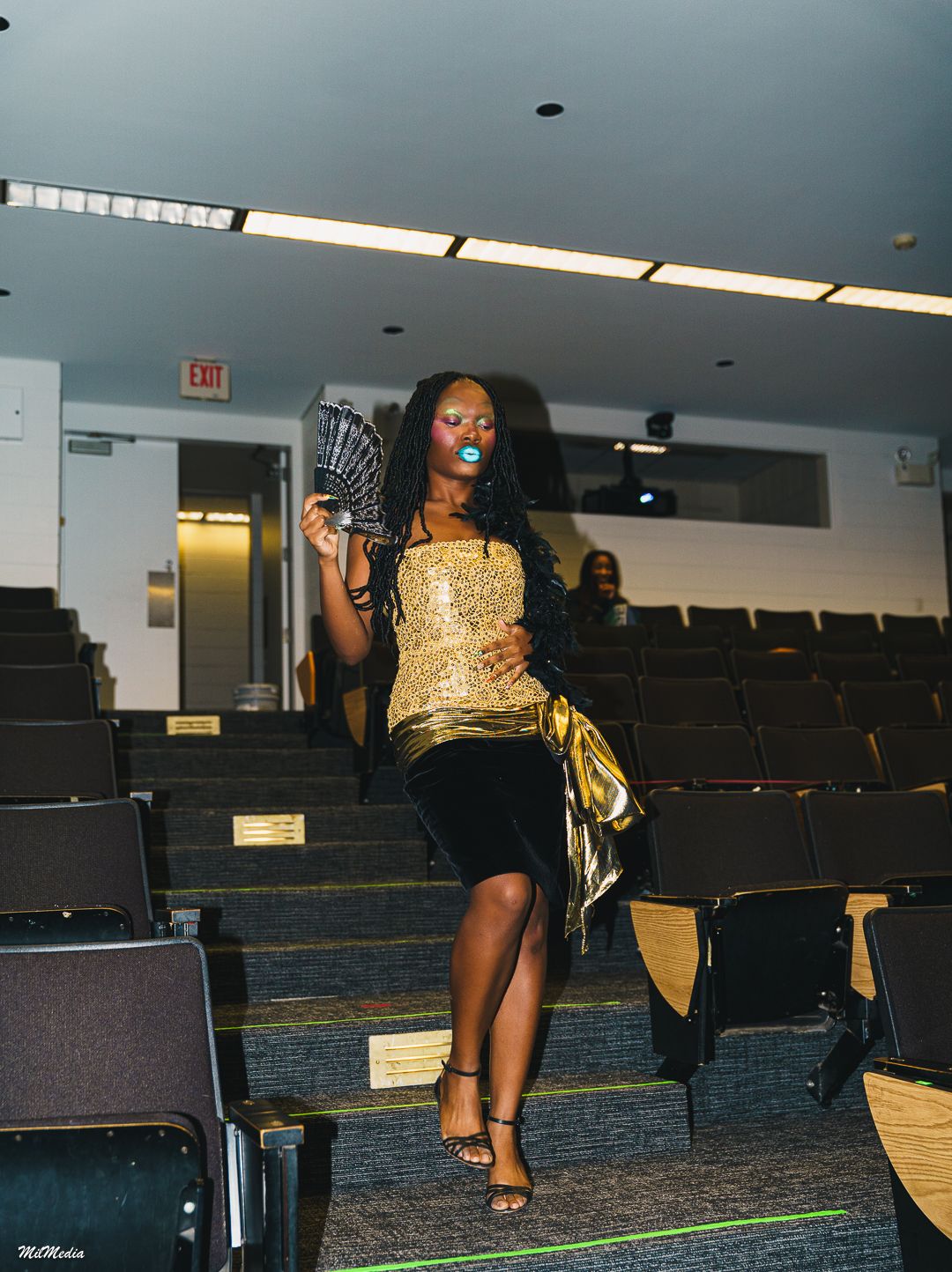 A woman in a gold sequin top and black skirt holds a fan while walking through an auditorium.