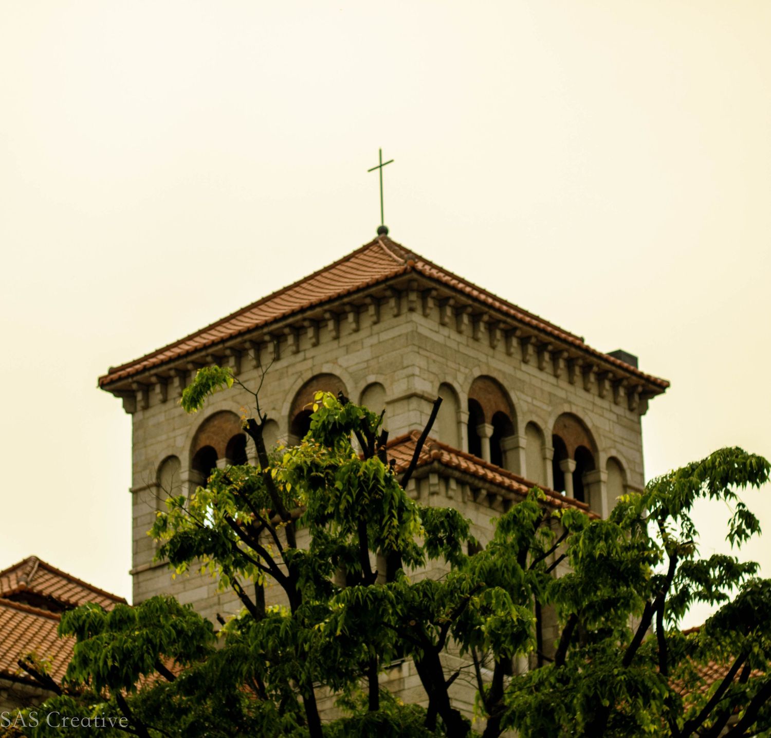 Stone church bell tower with a cross rising above green trees against a pale sky.
