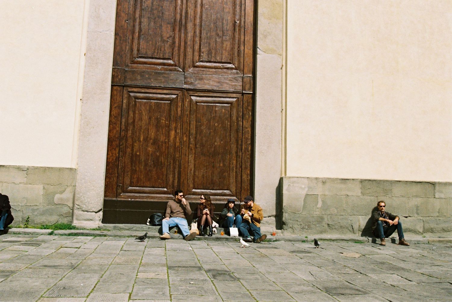 Group of people sitting and resting against a large wooden door on a stone plaza.