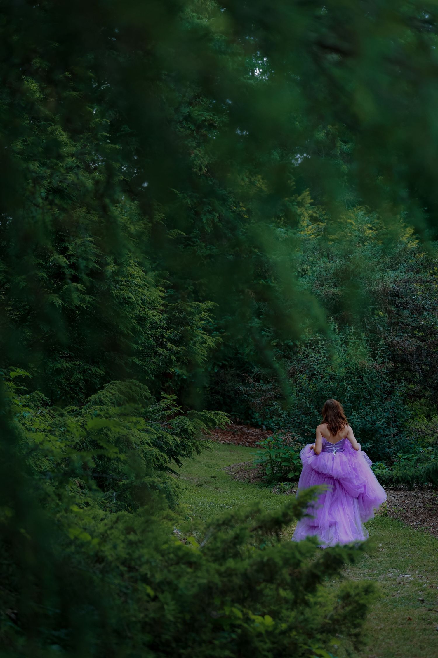 Woman in purple tulle gown walking through lush green garden path surrounded by dense trees.
