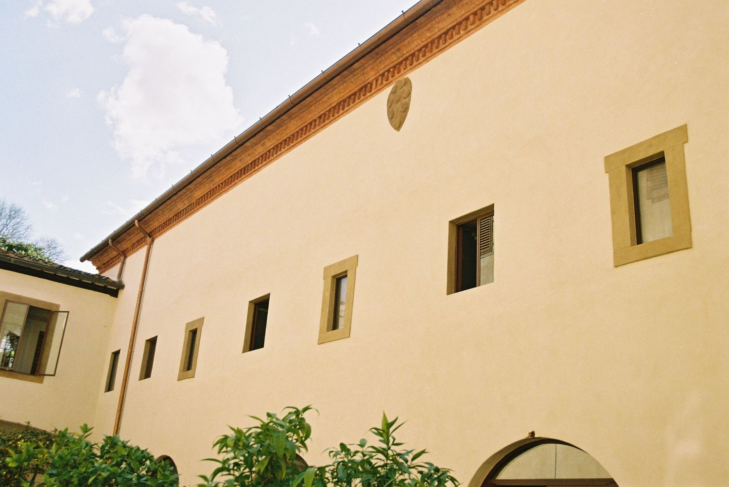 A sunlit Italian Renaissance building with cream-colored walls, terracotta cornice, and arched doorway surrounded by lush greenery.