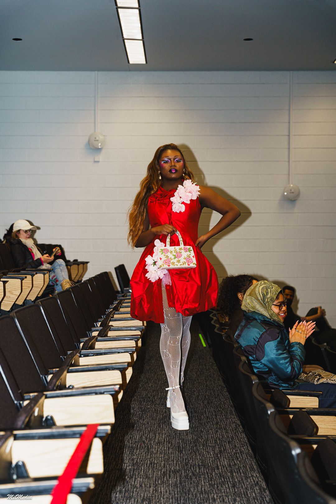 Model walks runway in red bubble dress with white floral accents, white tights, and platform shoes at indoor fashion show.