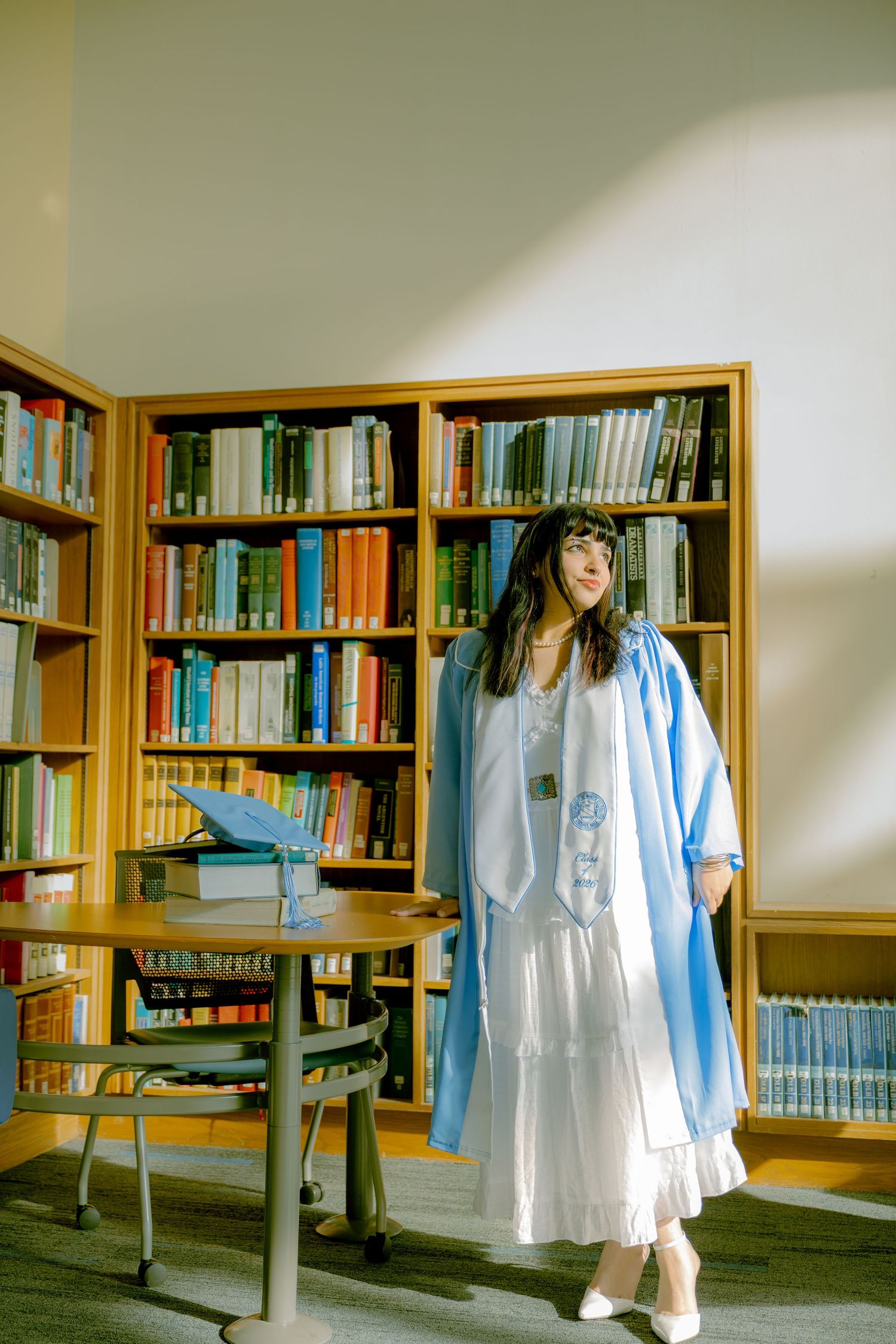 Graduate in blue gown and white dress poses in a sunlit library surrounded by bookshelves.