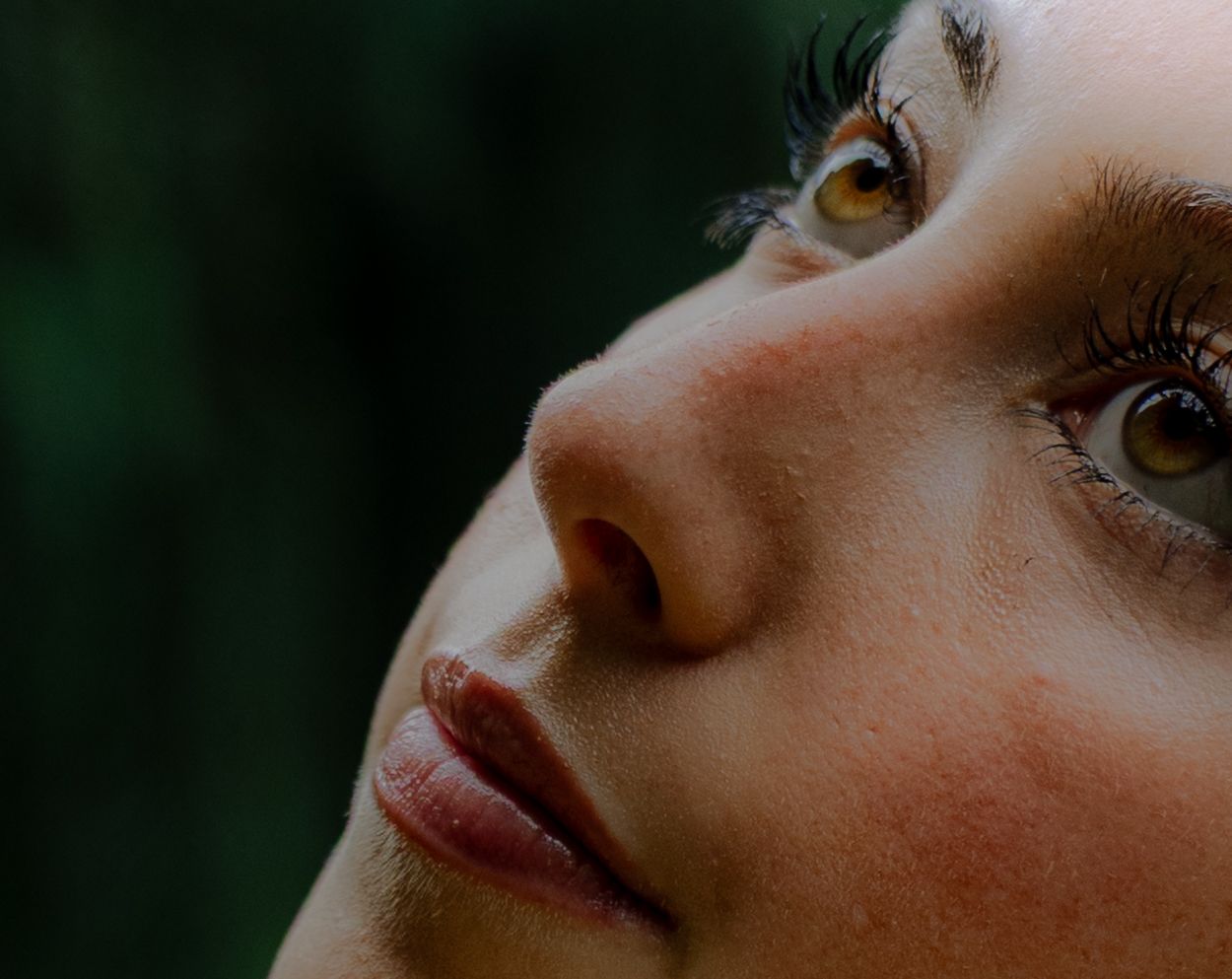 Close-up portrait of a young woman with hazel eyes and dark lashes gazing upward against a blurred green background.