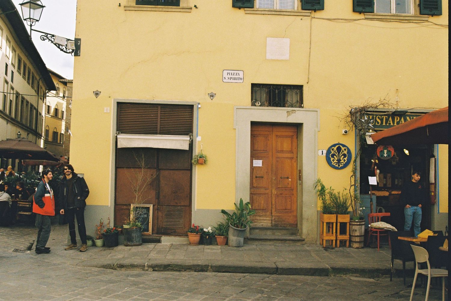 Pedestrians walk past a yellow corner building with a wooden door and adjacent restaurant on a cobblestone street in Italy.
