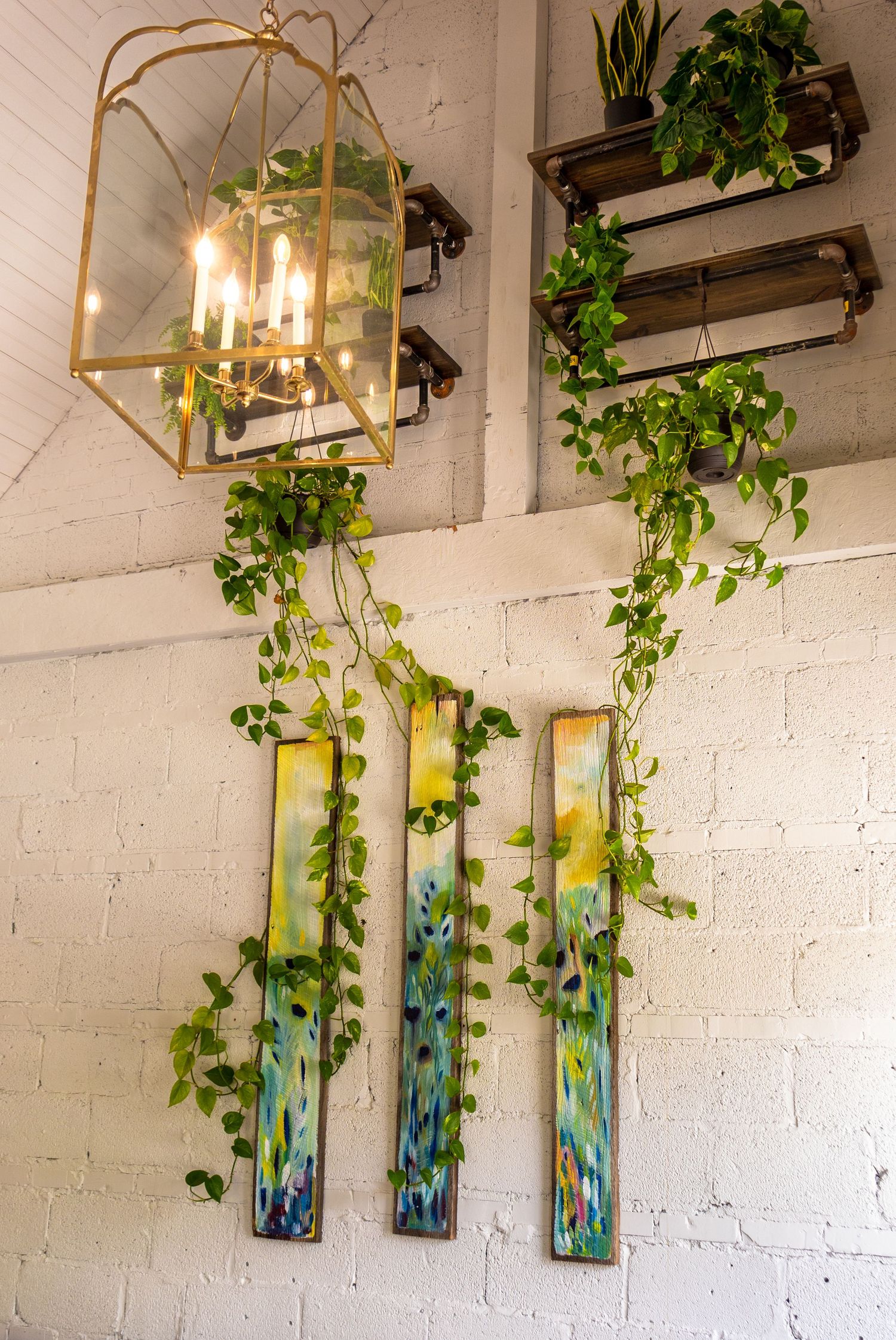 White brick wall cafe interior with gold lantern chandelier, trailing pothos vines, and colorful abstract wall art panels.