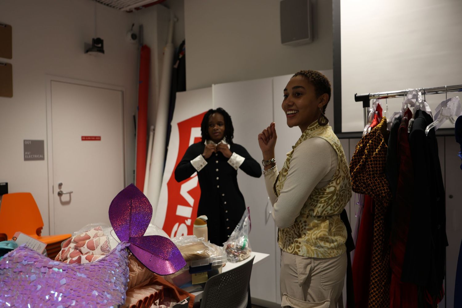 Two women in a backstage dressing room surrounded by colorful costumes and accessories before a show.