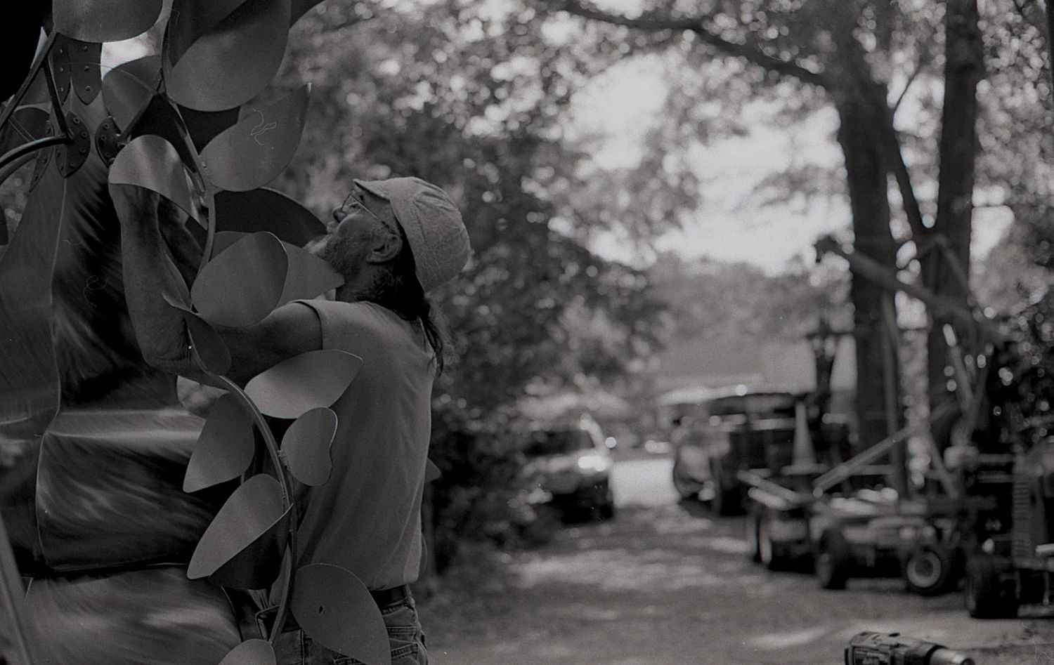 Black and white photo of a person resting beside a metal leaf sculpture with vehicles in background.