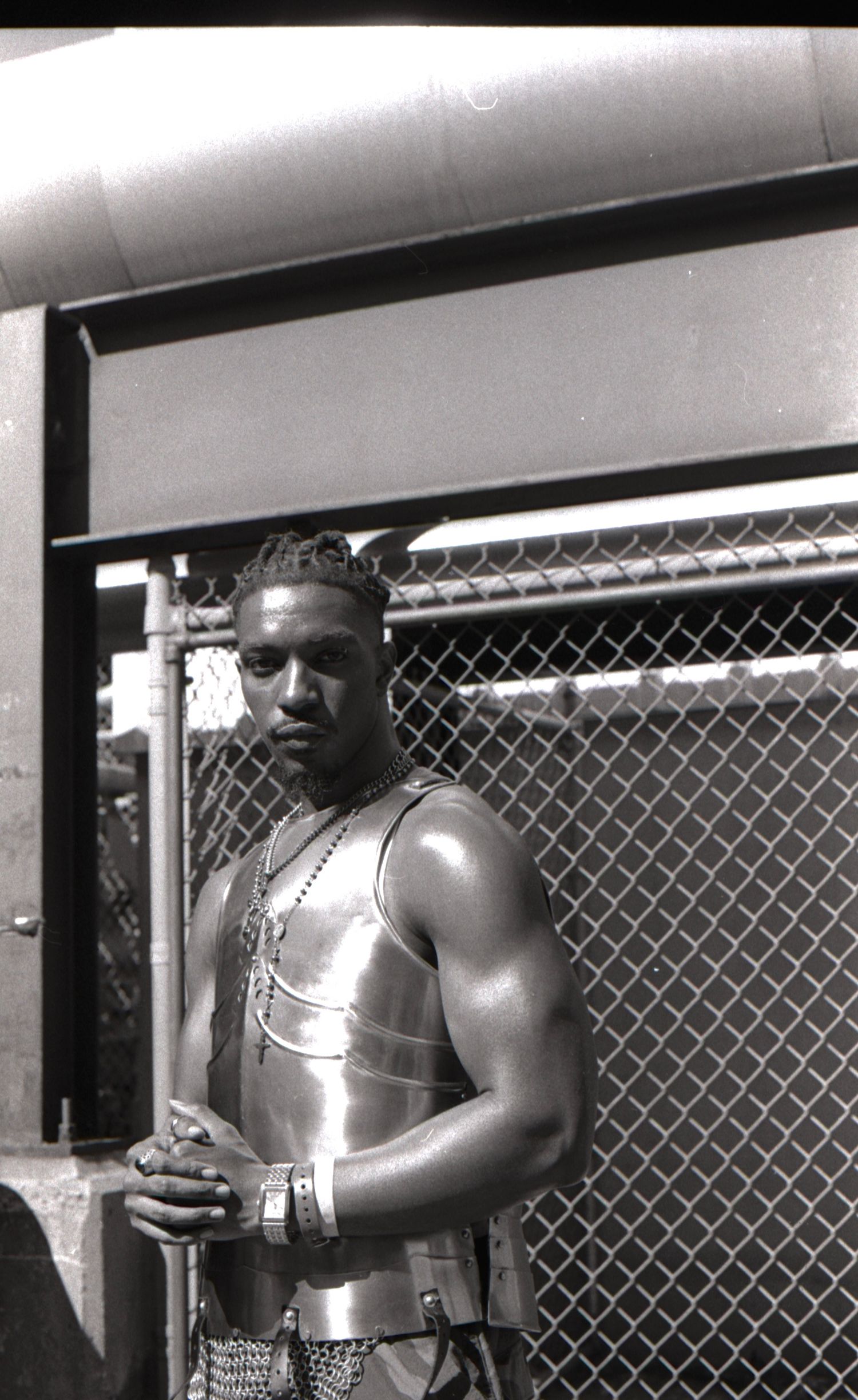 Black and white portrait of a man in metallic armor vest leaning against a chain-link fence.
