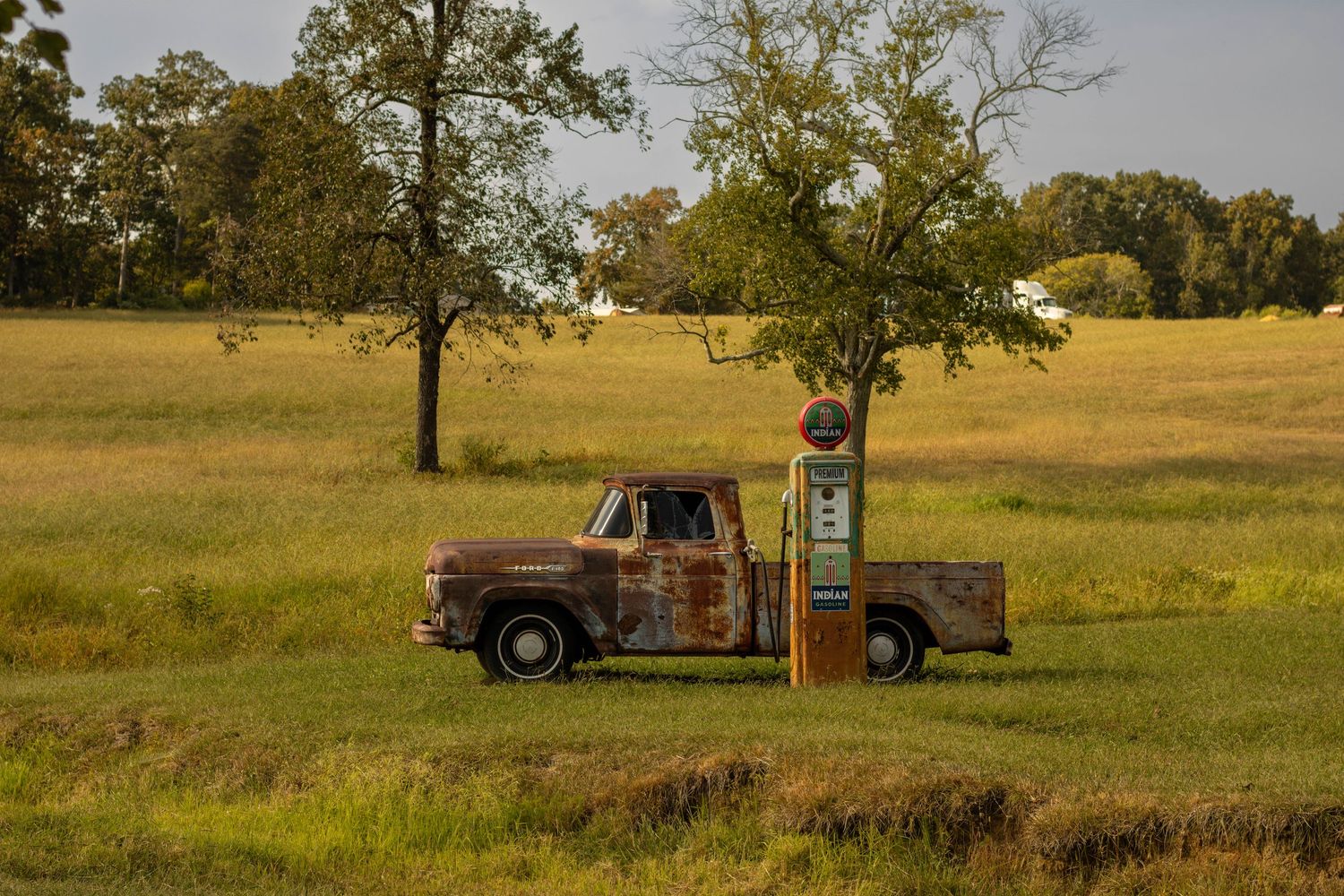 Rusty vintage pickup truck parked beside an old gas pump in a green rural field.
