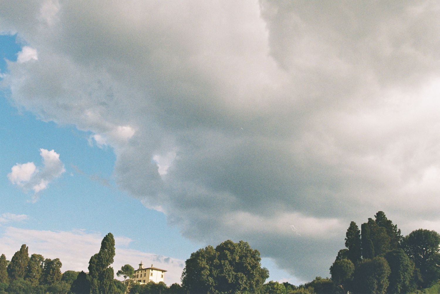 Dramatic cloudy sky over lush green trees and a yellow building in the distance.