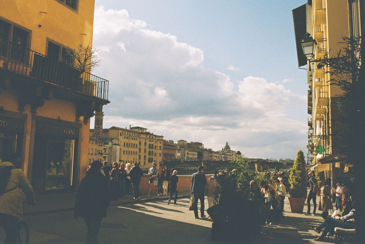 Crowds of tourists gather on a sunny piazza in Florence, Italy, with historic buildings and cloudy sky.
