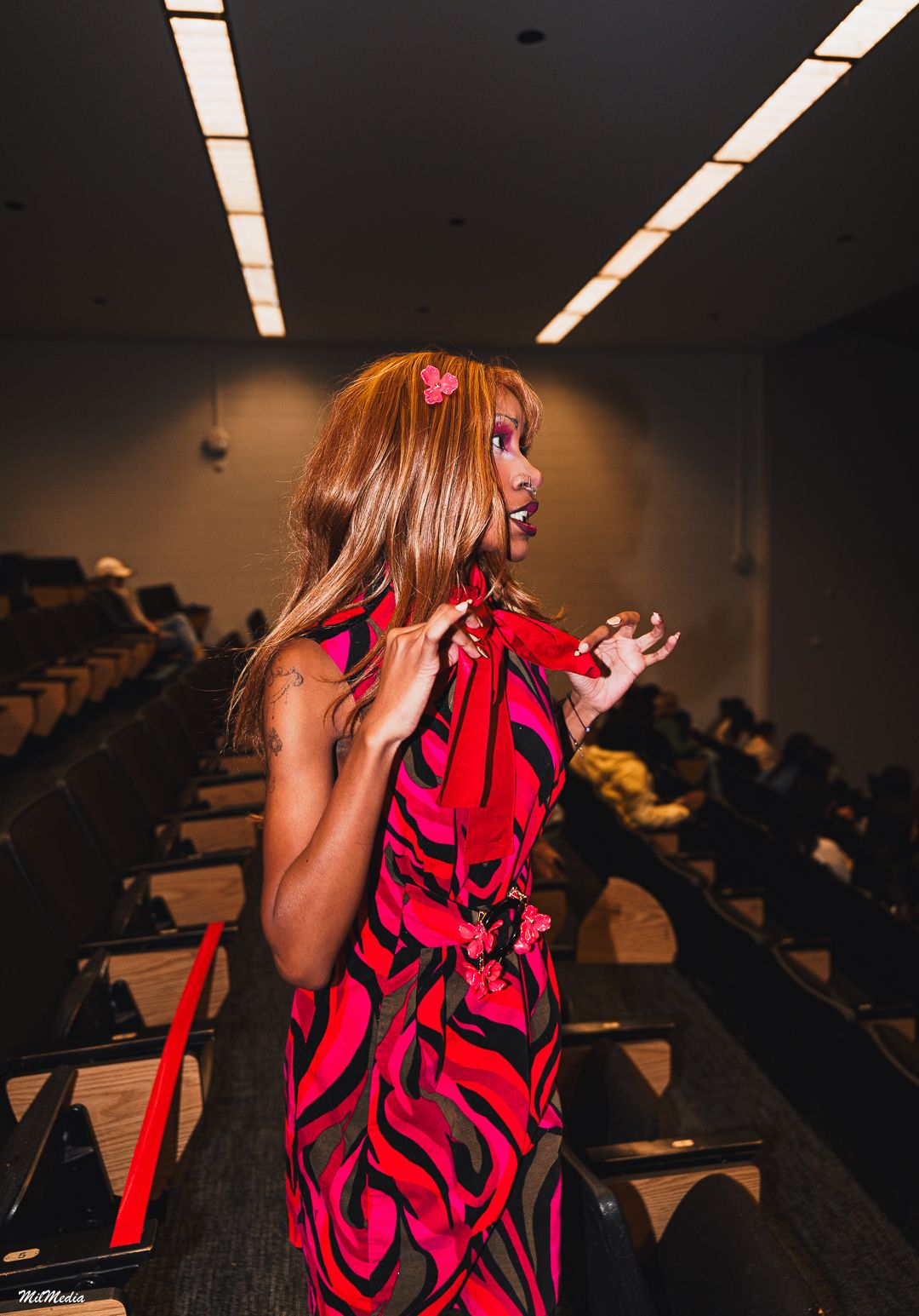 A woman in a bold red and pink patterned dress stands in a lecture hall aisle.
